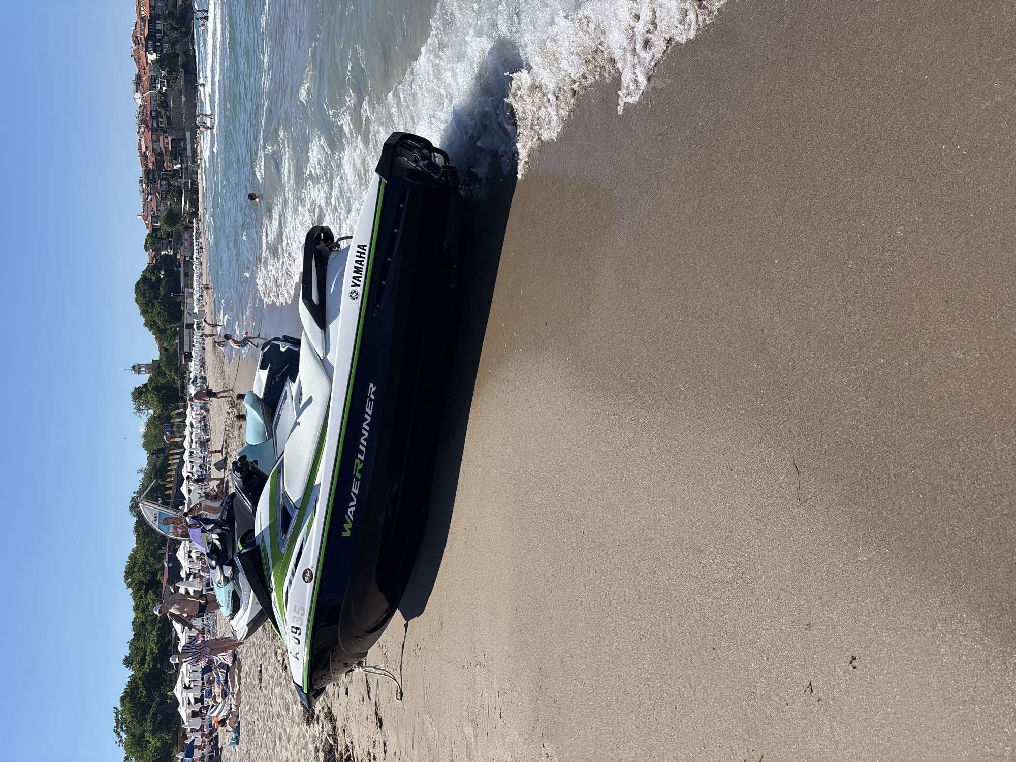 This media file depicts a busy beach scene on a sunny day. The primary subjects are several jet skis parked on the sandy shore, with one in the foreground partially in the water. The jet ski in the foreground is a Yamaha WaveRunner, white and green, with the model name "WAVERUNNER" visible in green text. There is also visible text "YAMAHA" on the jet ski. The background shows a lively beach with many people sunbathing on lounge chairs and swimming in the ocean. The water is clear and has small waves rolling onto the shore. Behind the beach, there are trees and buildings that suggest a coastal town or resort. The sky is a clear blue, indicating fair weather. The overall impression is one of a recreational summer day at the beach.