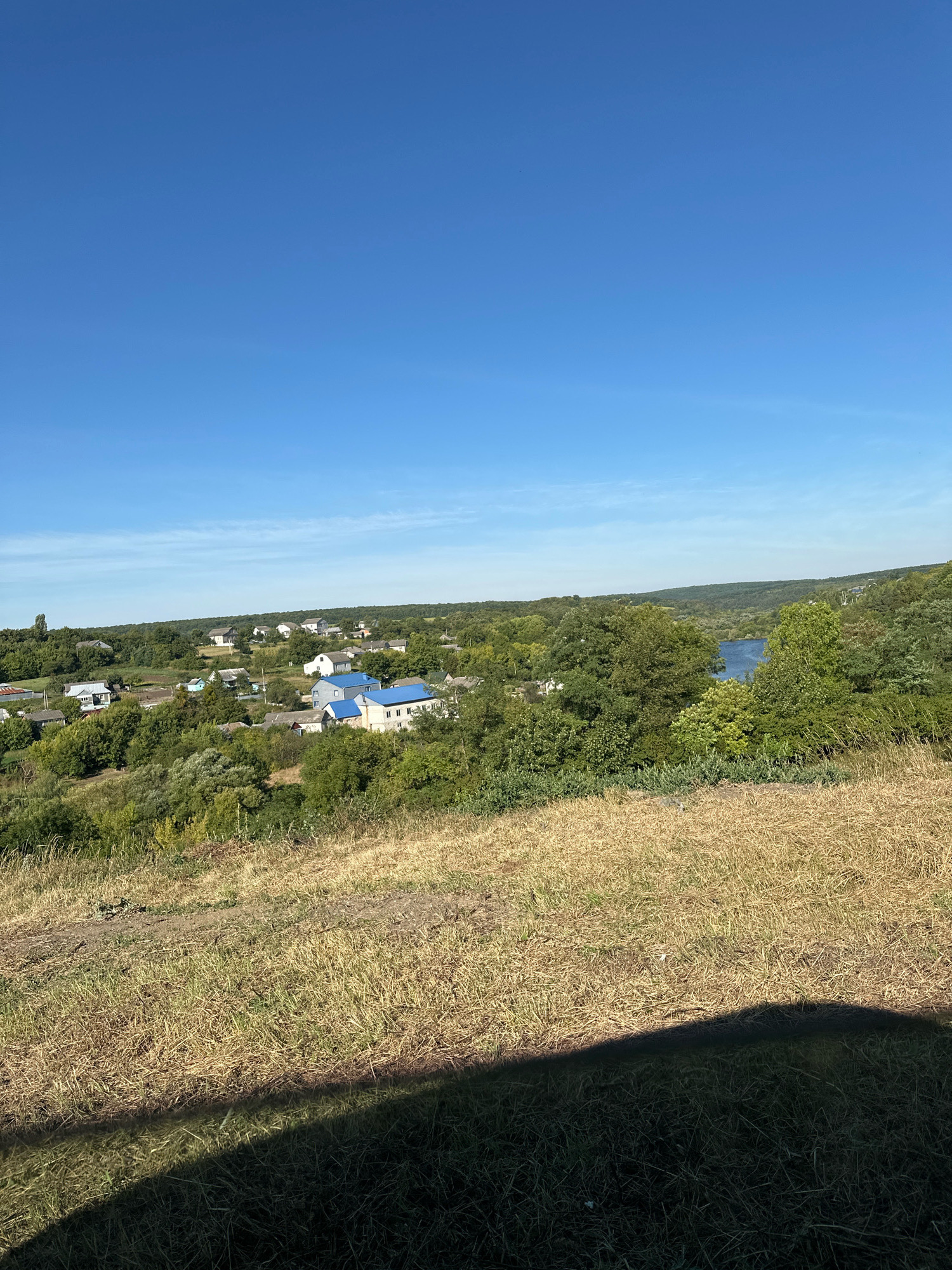 This is a wide shot of a rural landscape under a clear blue sky. In the foreground, dry, yellowed grass covers a sloping hill. A dark shadow, likely from a person or object outside the frame, stretches across the bottom right corner of the image.
In the midground, a village is nestled among lush green trees. Several houses with white and blue roofs are visible. A winding river or large body of water can be seen in the distance, reflecting the blue sky. The background consists of rolling hills covered in dense vegetation. The scene appears to be a quiet, natural setting, possibly on the outskirts of the village of Makiv, Ukraine, given the style of the houses and the surrounding landscape which is characteristic of Ukrainian rural areas. There are no people or animals visible in the image.