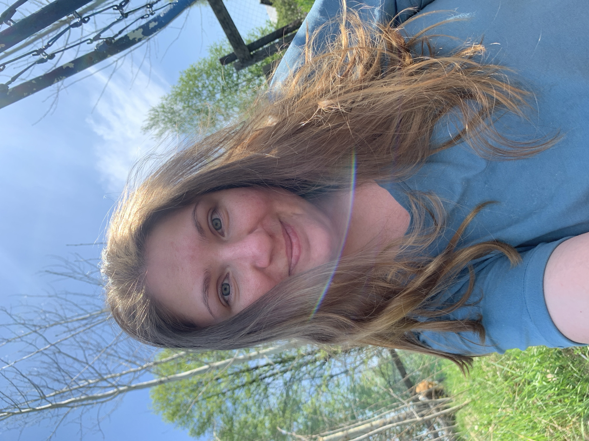 The image shows a person with long, light brown hair and fair skin. They are wearing a blue shirt. The background includes a blue sky with thin clouds, bare tree branches, green trees with leaves, and green grass. A metal fence is visible in the top left corner. The presence of the natural environment and the fence suggest the location might be a park or outdoor recreational area, possibly in a semi-urban setting. It's not possible to confirm it is in Zasechnyi, Russia, or any other specific location, based on visual information alone.