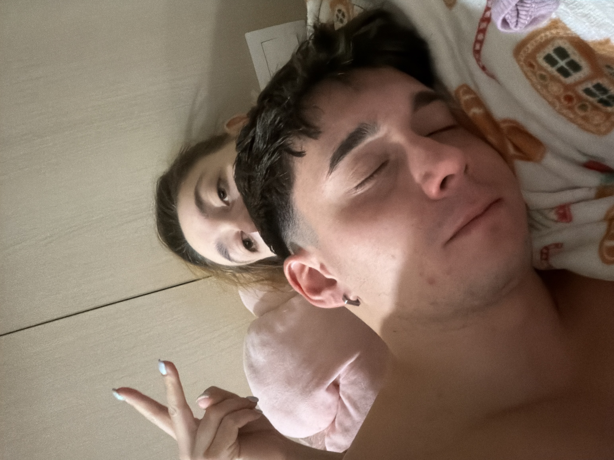 This image depicts an indoor bedroom scene in Sofia, Bulgaria, featuring two individuals in a bed. In the foreground, a male individual is visible from the upper torso, lying on his side with his eyes closed, appearing to be asleep. He has short, dark hair and a dark, geometric-shaped earring in his visible left ear. His bare shoulder is prominent. His head rests on a white pillow patterned with gingerbread houses and other festive designs. Behind and partially obscured by the male, a female individual is visible. Her left eye is open and directed towards the camera. She has light brown hair. Her left hand is raised in a 'V' or peace sign gesture, with fingernails painted light blue. A pink fabric, likely a blanket or clothing, is partially visible around her. The background consists of a plain, light-colored wall with a distinct vertical seam and a portion of a light fixture visible on the upper right side of the frame.