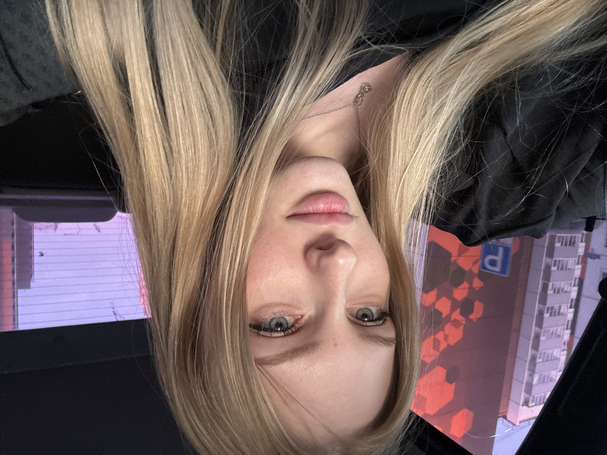 The image presents a young woman with blonde hair and green eyes, potentially in her late teens or early twenties. She is wearing a black top and a silver necklace with an infinity symbol. The background shows a reflection of buildings, including one with a prominent "P" sign, indicating a parking structure. The location is identified as Kondratovo, Russia, based on the distinctive "P" signage style prevalent in the region. The scene appears to be captured from inside a vehicle, possibly a car, suggesting the woman is either a passenger or the driver capturing a selfie.