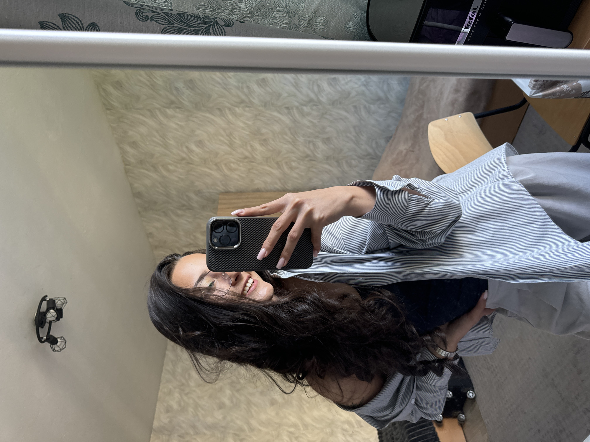 A woman is taking a selfie in a mirror indoors. She is holding a phone with a dark case and smiling. The woman has long, dark, wavy hair, is wearing glasses and a striped shirt. A light fixture is visible on the wall behind her on the left. A table and a small wooden chair can be seen on the right side. The background wallpaper has a subtle, swirling pattern. A "Tefal" appliance is visible on the table. The location is potentially an apartment or home within Sofia, Bulgaria.