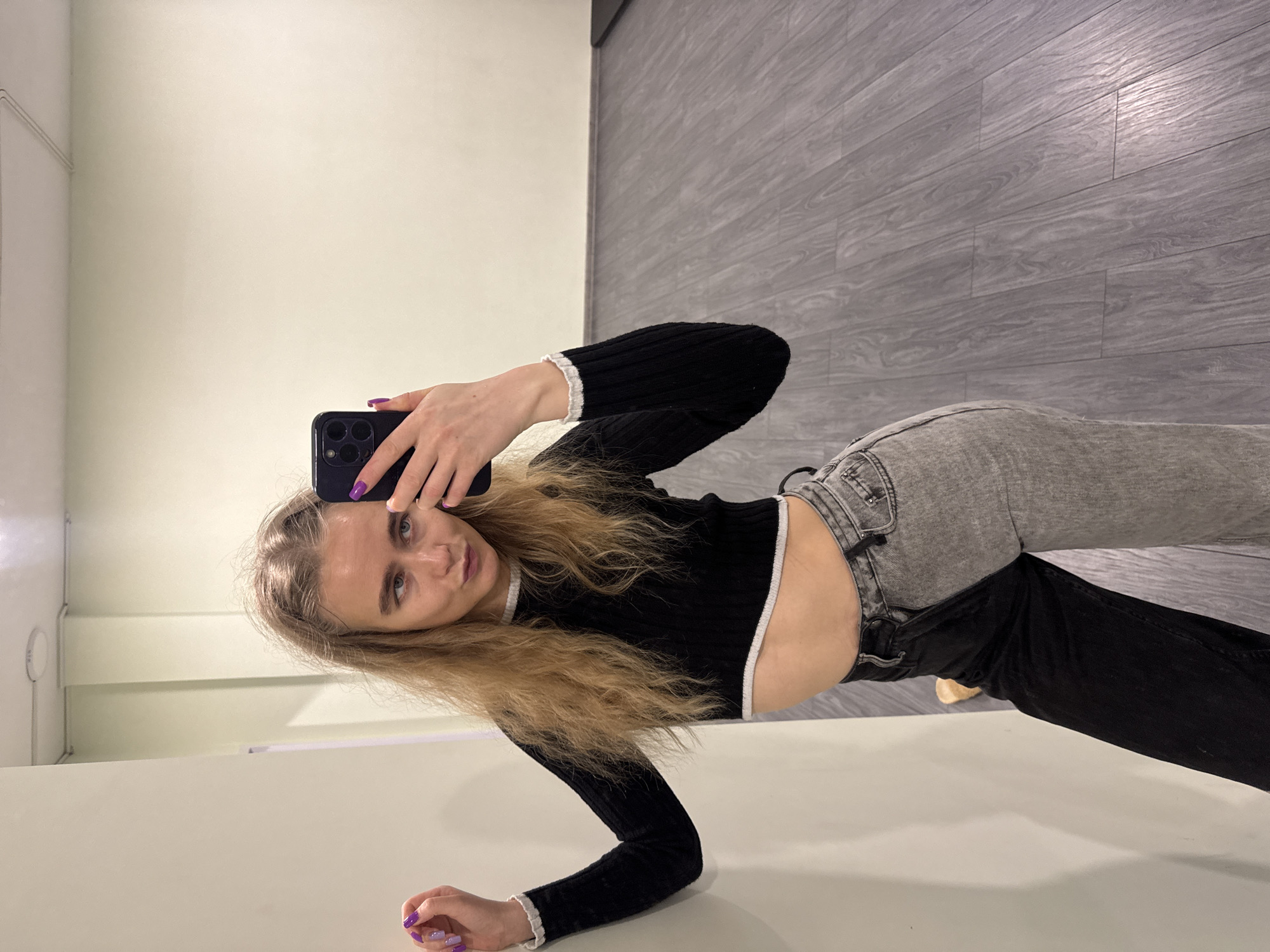 A young woman is indoors, possibly in a clothing store fitting room, taking a selfie with a black smartphone. She is leaning on a white surface, possibly a table or bench. She is wearing a black, long-sleeved, ribbed crop top with white trim and two-tone jeans that are gray on top and black on the bottom. The background shows a light beige wall and gray laminate flooring. Her nails are painted purple. The location is Unknown, Unknown.