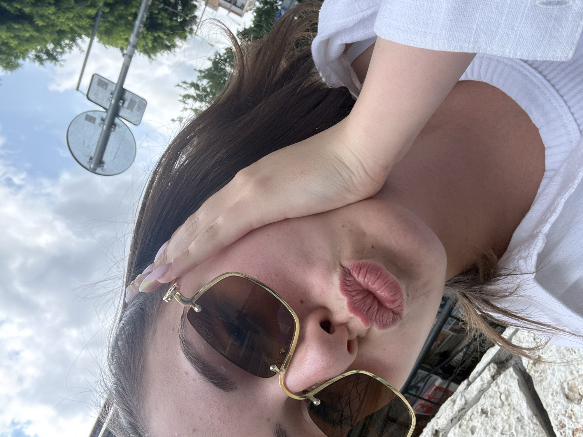 In a self-portrait taken outdoors, a young woman with long brown hair and fair skin leans her right cheek against her hand, her lips pursed in a kissing gesture. She is wearing gold-rimmed sunglasses with brown lenses, featuring the "GUCCI" logo on the left lens. Her fingernails appear to be painted with a light, pastel color. She is dressed in a white collared shirt with the collar visible. The background suggests an urban setting with a bright, cloudy sky above, trees, and a street signpost. The lighting indicates it is daytime. There is no specific indicator within the image to place the location in the city of Burgas, Bulgaria.
