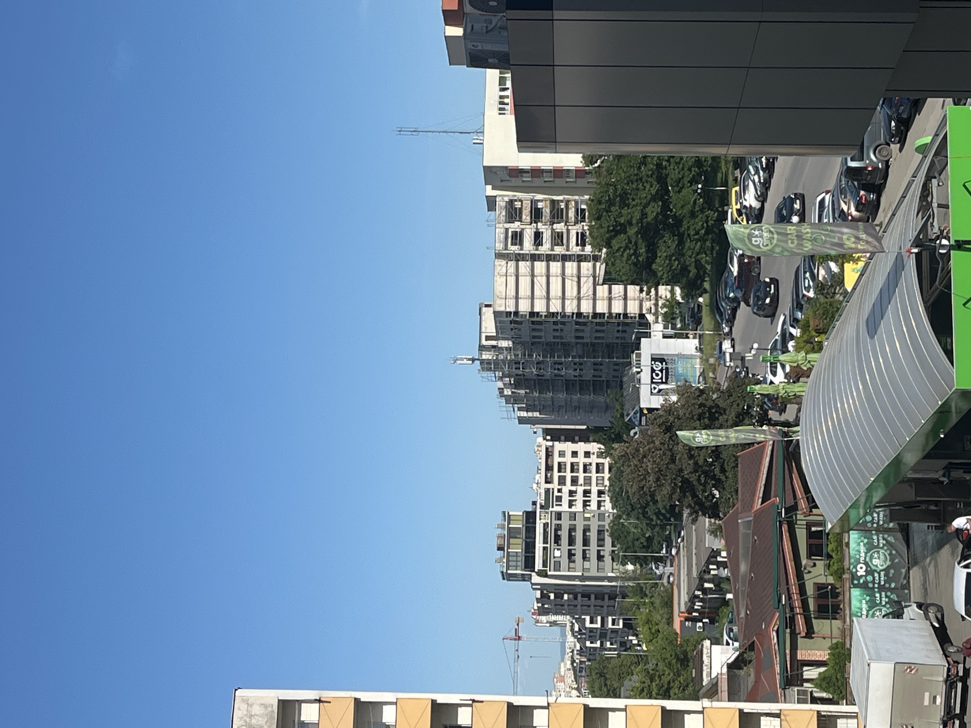 A medium, eye-level shot shows a cityscape under a clear blue sky. The focus is on the buildings and structures in the urban environment. A variety of architectural styles are visible, from older buildings with sloping roofs to modern high-rises under construction. Some buildings are undergoing construction with scaffolding around them. A crane is visible in the distance, indicating ongoing development. The street is lined with parked cars and vegetation, suggesting a bustling urban area. There are also advertisements related to "Car Wash". The location appears to be in a city, but the specific location, including the city and country, is Unknown.
