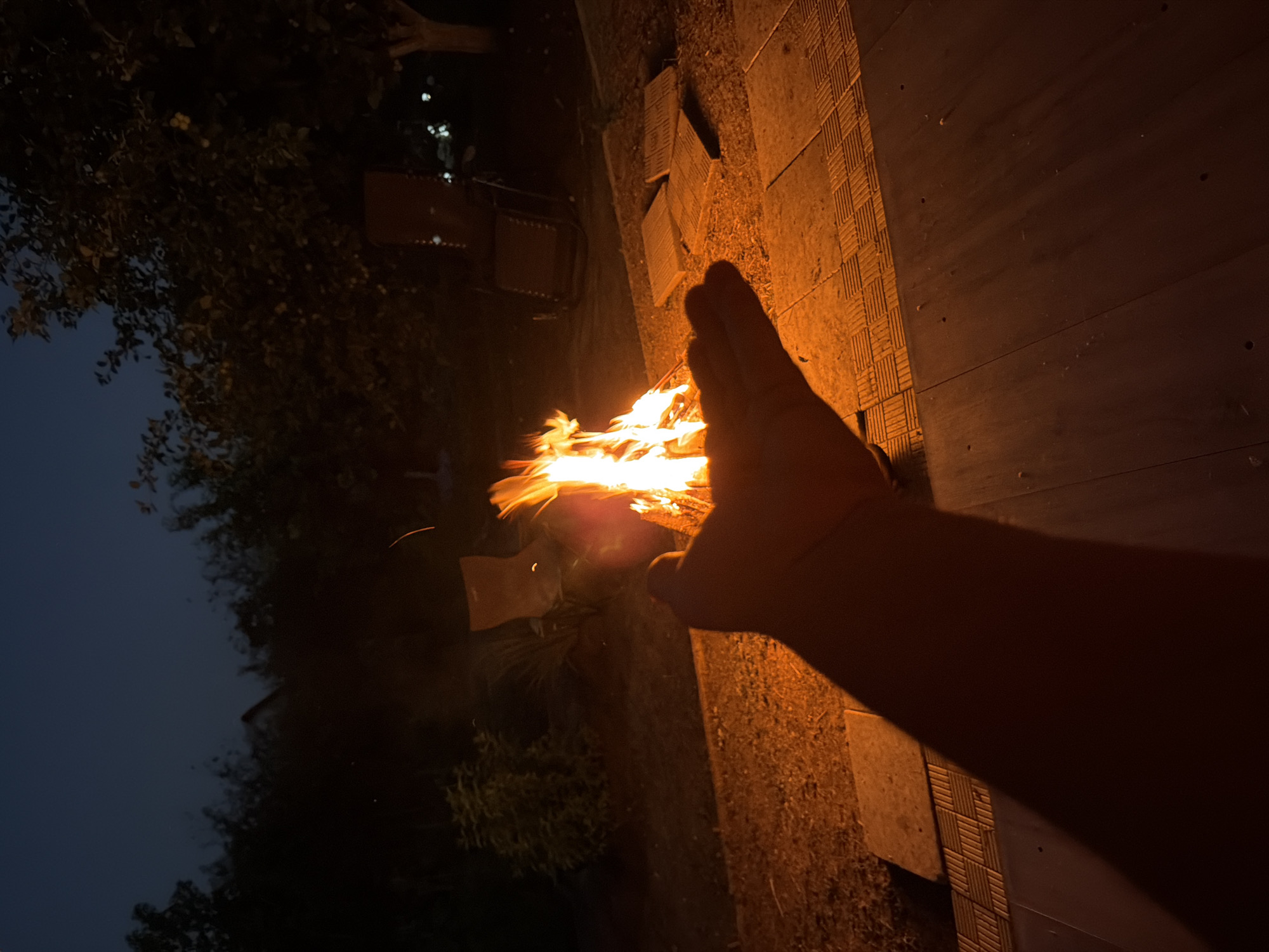 In Nezhinka, Russia, at dusk, a bonfire burns in a backyard, illuminating the surrounding area with a warm orange glow. The flames flicker and dance, casting shadows on the ground. In the foreground, a person's hand is extended towards the fire, seemingly reaching out to feel its warmth. The hand is silhouetted against the bright flames, creating a dramatic effect.
The backyard is furnished with two lawn chairs, suggesting a space designed for relaxation and outdoor gatherings. The ground is covered with gravel and paved with square tiles, creating a rustic and inviting atmosphere. In the background, trees and bushes surround the area, adding to the natural ambiance. The sky is a deep blue, indicating the transition from day to night. The overall scene evokes a sense of peace and tranquility, with the bonfire serving as the central focal point, drawing attention to its mesmerizing flames and the comfort it provides.