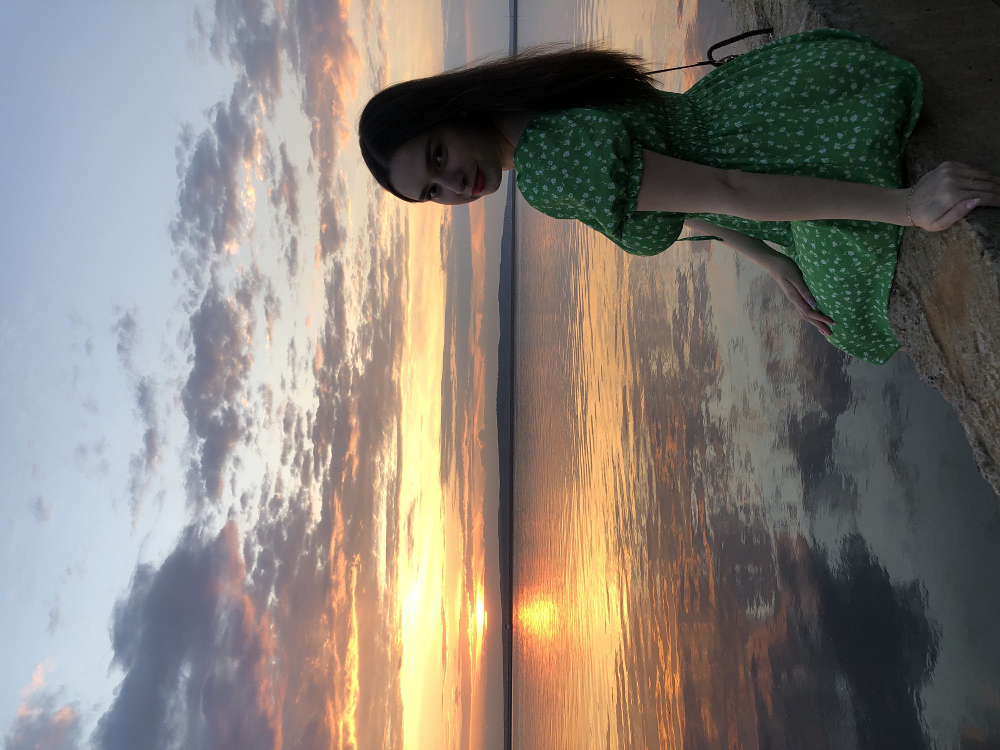 A woman in a green dress sits on a concrete structure overlooking a body of water at sunset. The sky displays a mix of blue, white, and orange-tinged clouds, reflected in the water. The sun is setting on the horizon, creating a bright glow. The woman has long, dark hair and is looking towards the camera. The location is likely in or near Chaikovsky, Russia, due to the provided context.
