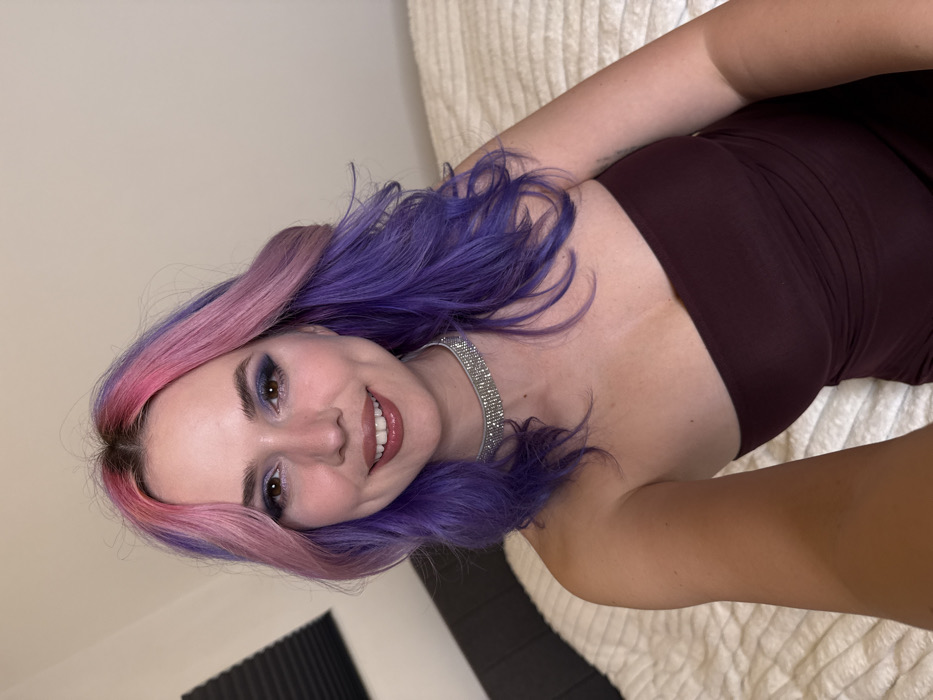 A female subject is depicted indoors, framed from the mid-torso up, appearing to take a selfie. She has fair skin, wavy hair dyed pink at the roots blending into purple ends, and is smiling directly at the camera. Her eyes are accented with dark blue or purple eyeshadow, and her lips are painted with a glossy reddish-brown lipstick. She wears a strapless, form-fitting maroon dress or top and a multi-row silver-toned rhinestone choker necklace. Her left arm is partially visible on the left side of the frame. The background consists of a light-colored, textured, possibly quilted blanket or comforter behind her, and a plain light beige wall. A dark headboard or furniture piece is partially visible on the left side, along with a dark, ridged object in the upper left corner. The specific location within the City of Zagreb, Croatia, cannot be determined from the visual information provided.