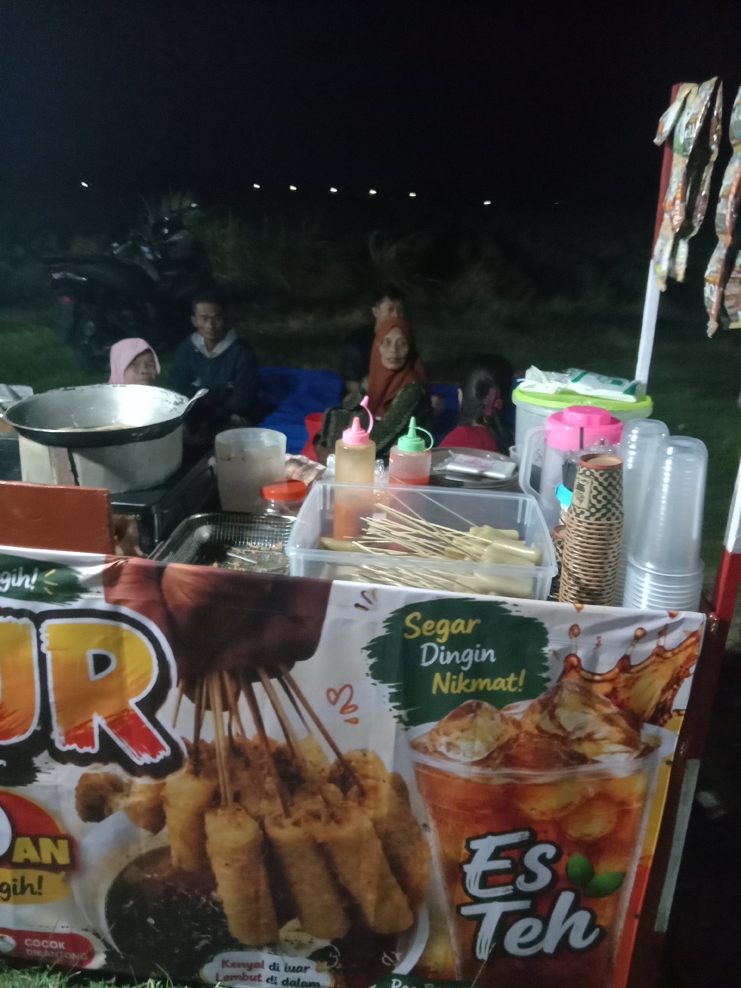 At night, a food stall is set up outdoors, likely in a street or open area in Stiaji, Indonesia. The stall features a variety of food items and drinks. A prominent banner on the stall displays text in Indonesian, including "Es Teh," indicating iced tea. To the left, a large wok sits on a stove, with fried food items on skewers being prepared or displayed. Several people, including adults and possibly children, are gathered around the stall, some seated on the ground, suggesting a casual, communal atmosphere. The lighting is dim, typical of an evening setting, with some artificial light from the stall and possibly distant lights in the background. The ground appears to be covered in grass.