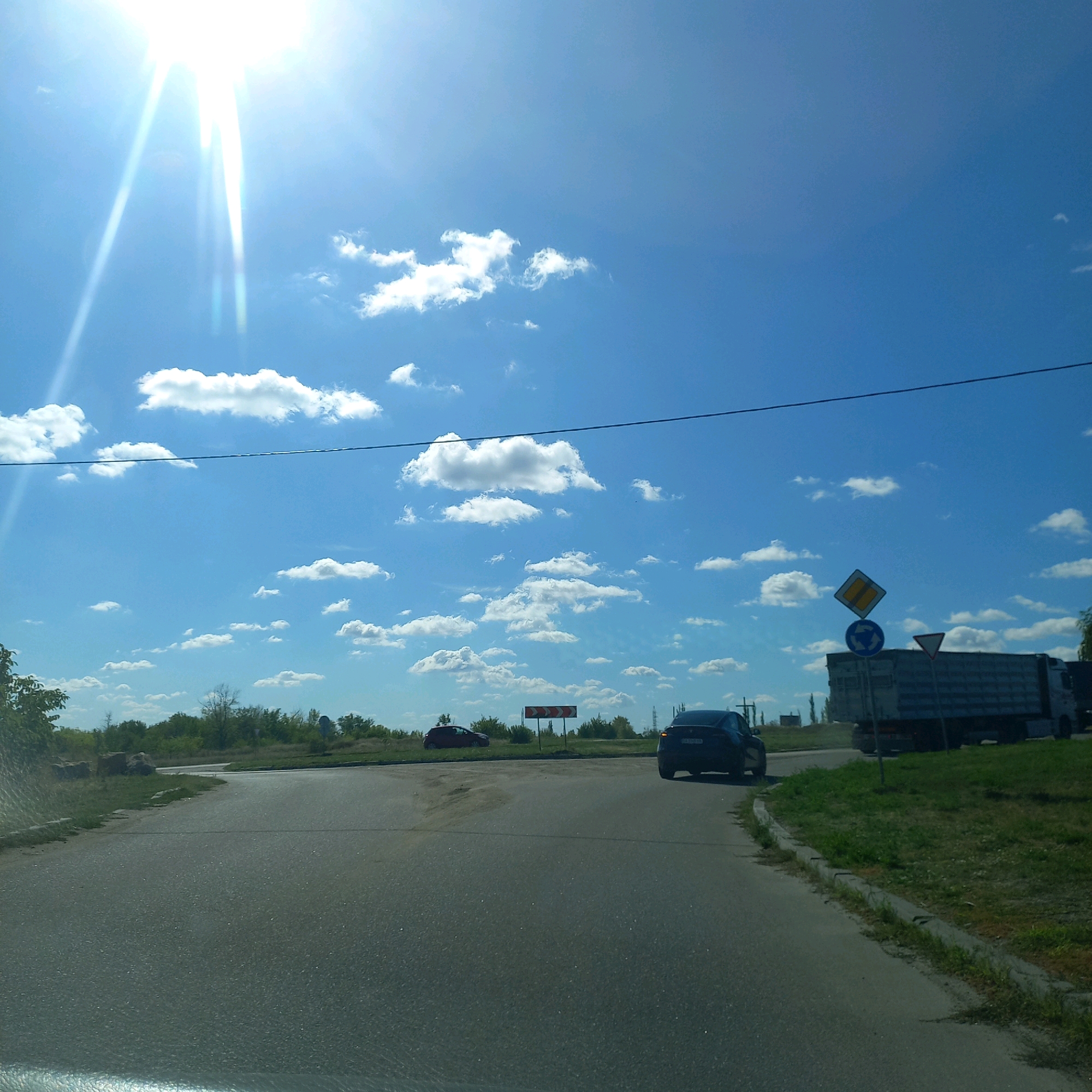 The image depicts a sunny day with a bright blue sky and scattered white clouds, likely taken from inside a vehicle. In the foreground, a paved road curves to the right. On the left side of the road, there is a patch of grass with some rocks and vegetation. In the middle distance, there is a road intersection with signage. A red car is visible on the left side of the intersection, and a dark-colored car is turning right in the foreground on the right side of the road. Further in the distance, a large truck is parked or moving, and more trees and vegetation line the horizon. The overall scene suggests an intersection in or near a rural or suburban area. The location is identified as Soniachne, Ukraine.