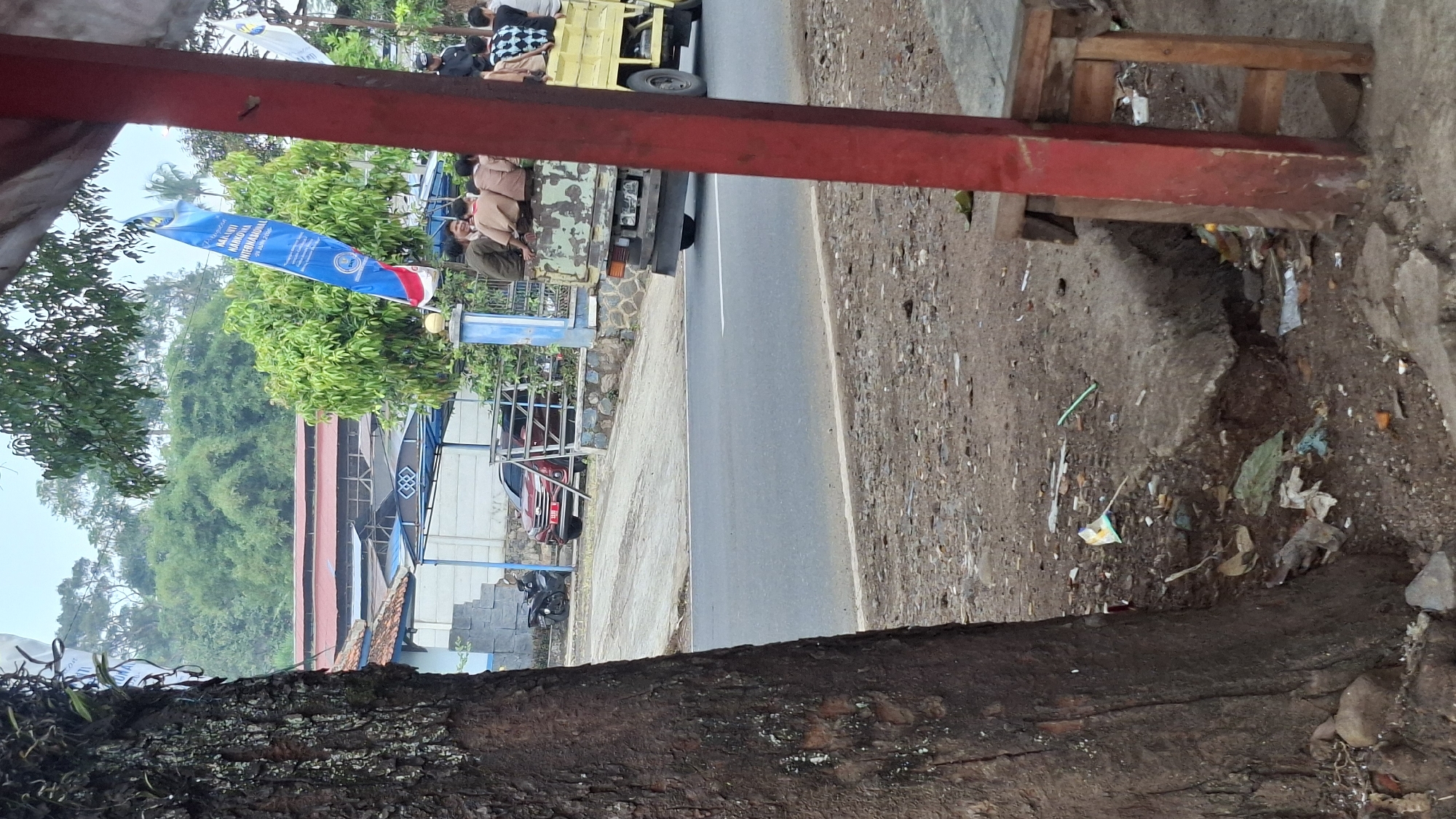 In Cilaku, Indonesia, a scene unfolds beside a paved road, suggesting a semi-urban or rural setting with abundant greenery. The foreground is dominated by the rough, textured bark of a large tree trunk on the left and the weathered, red-painted wooden post of a simple shelter on the right. Scattered debris, including plastic wrappers and what appear to be cigarette butts, litters the unpaved ground near the tree and under the shelter.
Across the road, a reddish-brown car is parked in front of a building with a red-tiled roof and a light-colored facade. A black motorcycle is parked nearby. Beyond the building, lush green trees and foliage cover the hillside, indicating the area's natural environment. A blue and white banner, seemingly promoting an anti-drug campaign with text in Indonesian, hangs from a pole.
A prominent object in the mid-ground is a flatbed truck, painted in faded green and yellow, carrying several young people, most of whom appear to be schoolchildren dressed in brown uniforms. They are seated in the back of the truck, facing towards the right side of the frame. The truck is in motion, moving away from the viewer and towards the right. A second truck, yellow, is visible further down the road. The sky is overcast, contributing to the subdued lighting of the scene.
