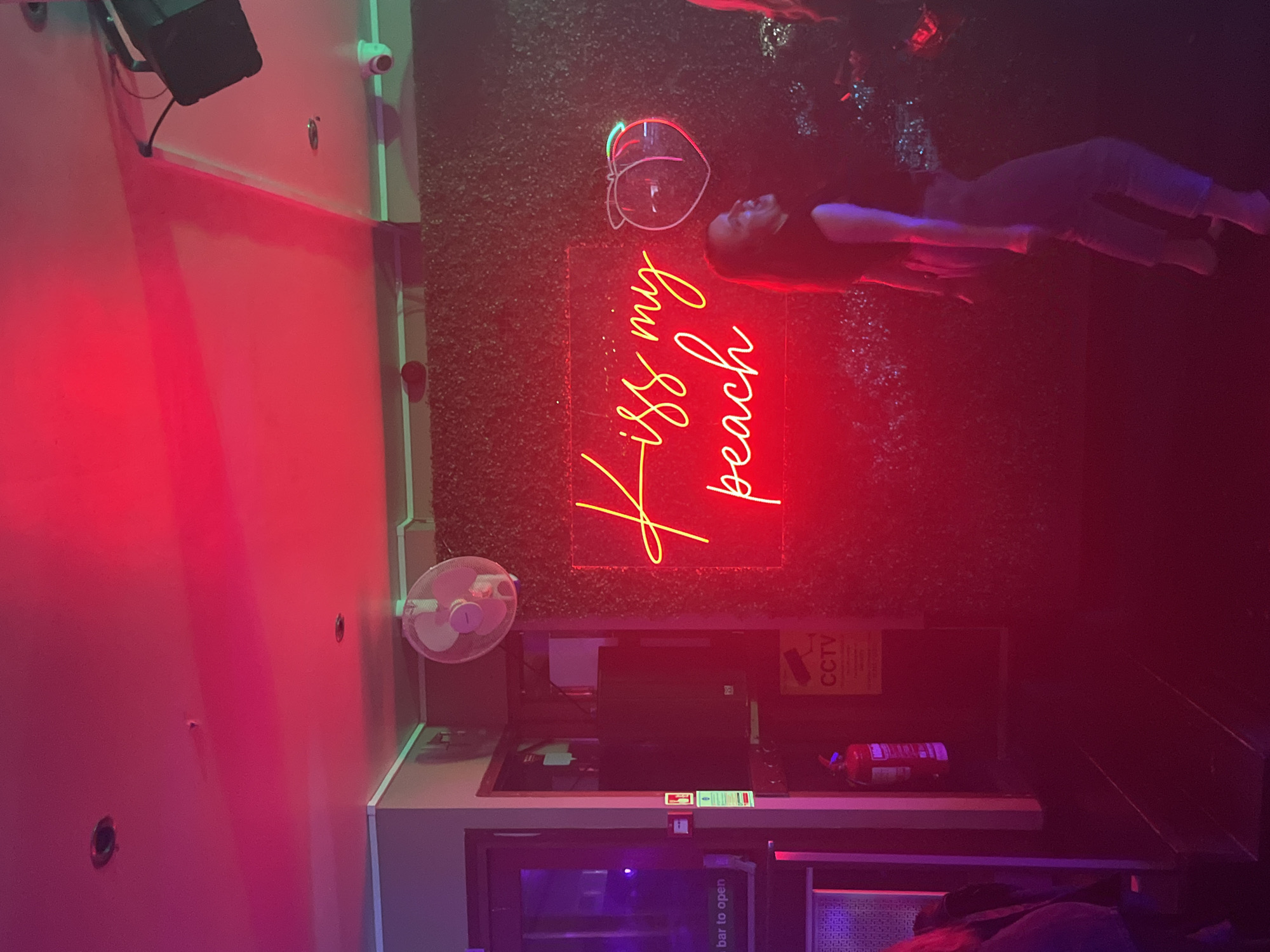The image shows the interior of a bar. A wall, painted red, is on the left. There are security cameras mounted on the wall, and a small electrical fan is also mounted on the wall.
There is a bar counter with a recessed sink. An item (possibly a fire extinguisher) is on top of the counter. A cooler appears under the bar counter. Above the bar counter, a neon sign says "Kiss my Peach", next to a peach. A woman is lying on her back. The location is likely to be in the city of Eastbourne, United Kingdom.