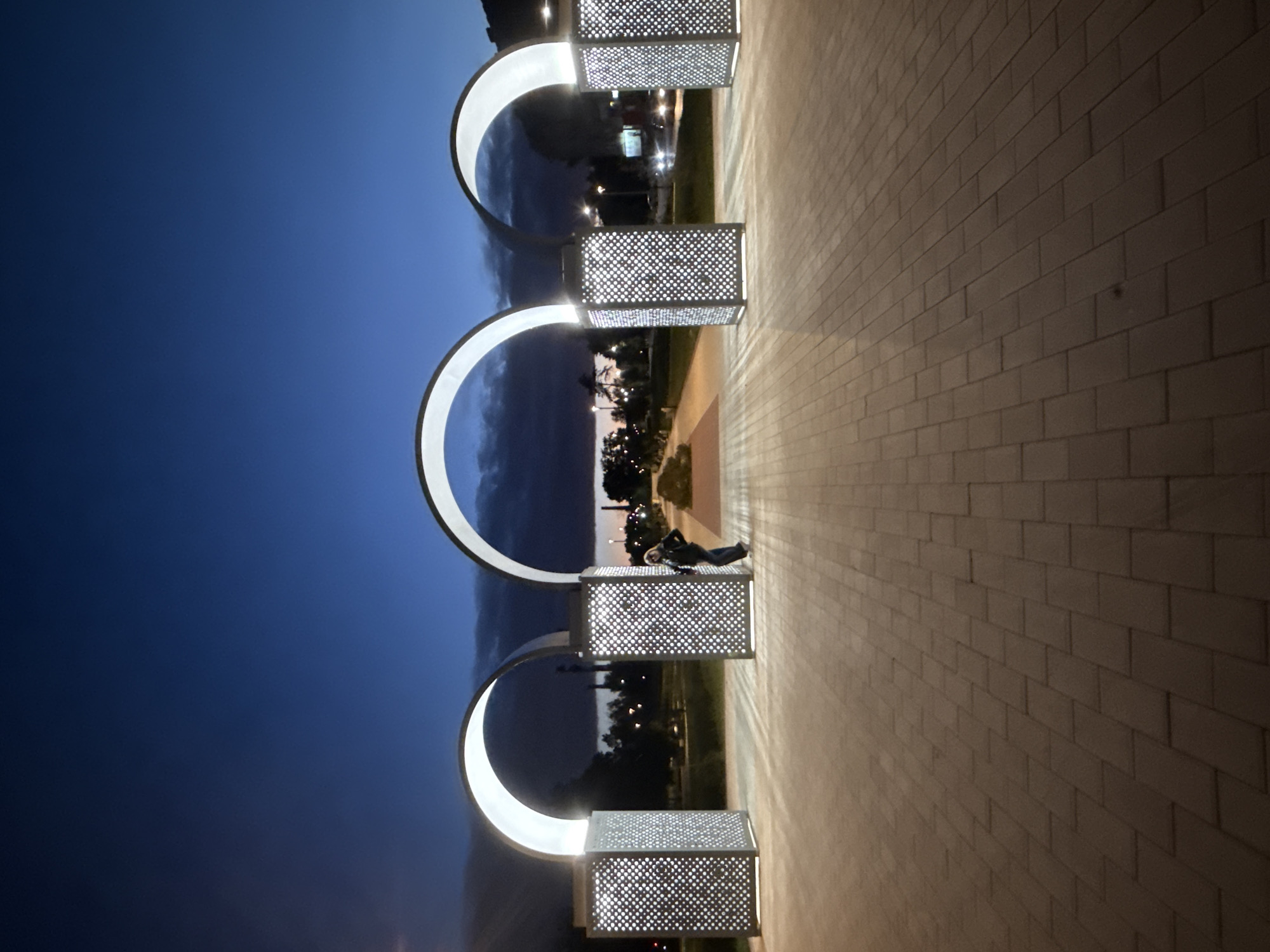 The image depicts an outdoor scene at dusk, likely in the city of Bor, Russia, judging by the provided context. Three illuminated archways, supported by patterned light columns, form a gateway or promenade. The pathway leading through the arches is paved with rectangular bricks. A single individual, wearing a dark jacket and pants and carrying a backpack, stands in the opening of the middle archway, facing away from the viewer and looking down the path. Beyond the arches, trees and streetlights are visible, indicating an urban or park setting. The sky is a deep blue, transitioning into darker tones at the horizon where clouds are present. The overall atmosphere is serene, with artificial lighting from the archways and distant streetlights creating a softly lit environment.