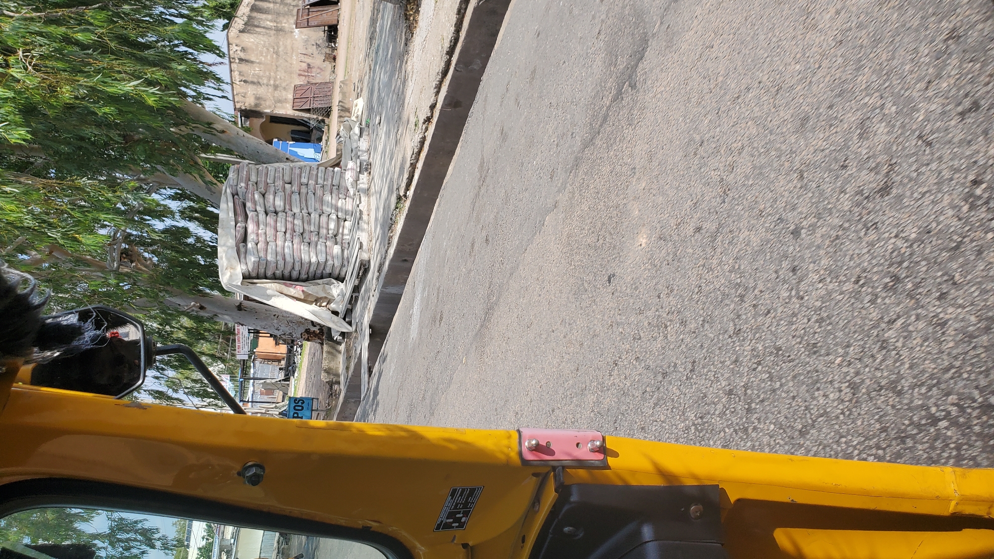 The image captures a street scene in Jalingo, Nigeria, from the perspective of someone inside a yellow motorized rickshaw (tuk-tuk). The rickshaw's bright yellow bodywork dominates the foreground, with a black side mirror visible on the left. Attached to the top of the rickshaw's body is a pink bracket with two silver screws.
To the left of the rickshaw, tall trees with green foliage cast shadows on the ground. Along the side of the road, a large stack of cement bags, wrapped in white plastic, is piled high. The bags appear to be uniform in size and color, suggesting they are all the same product.
Beyond the cement bags, there are buildings with weathered walls and metal doors, indicative of a commercial or residential area. A blue sign with white lettering is partially visible, and a smaller sign with "POS" is also discernible. The road surface is asphalt, with a concrete curb separating it from the area where the cement is stacked.
The lighting suggests it is daytime, with the sun casting strong shadows. The overall impression is of a bustling commercial area in a Nigerian city, with construction materials prominently displayed. The image is taken from a low angle, emphasizing the height of the cement bags and the surrounding trees. The orientation of the image is slightly tilted, creating a dynamic perspective. No people are clearly visible in the frame, focusing the viewer's attention on the objects and the environment.