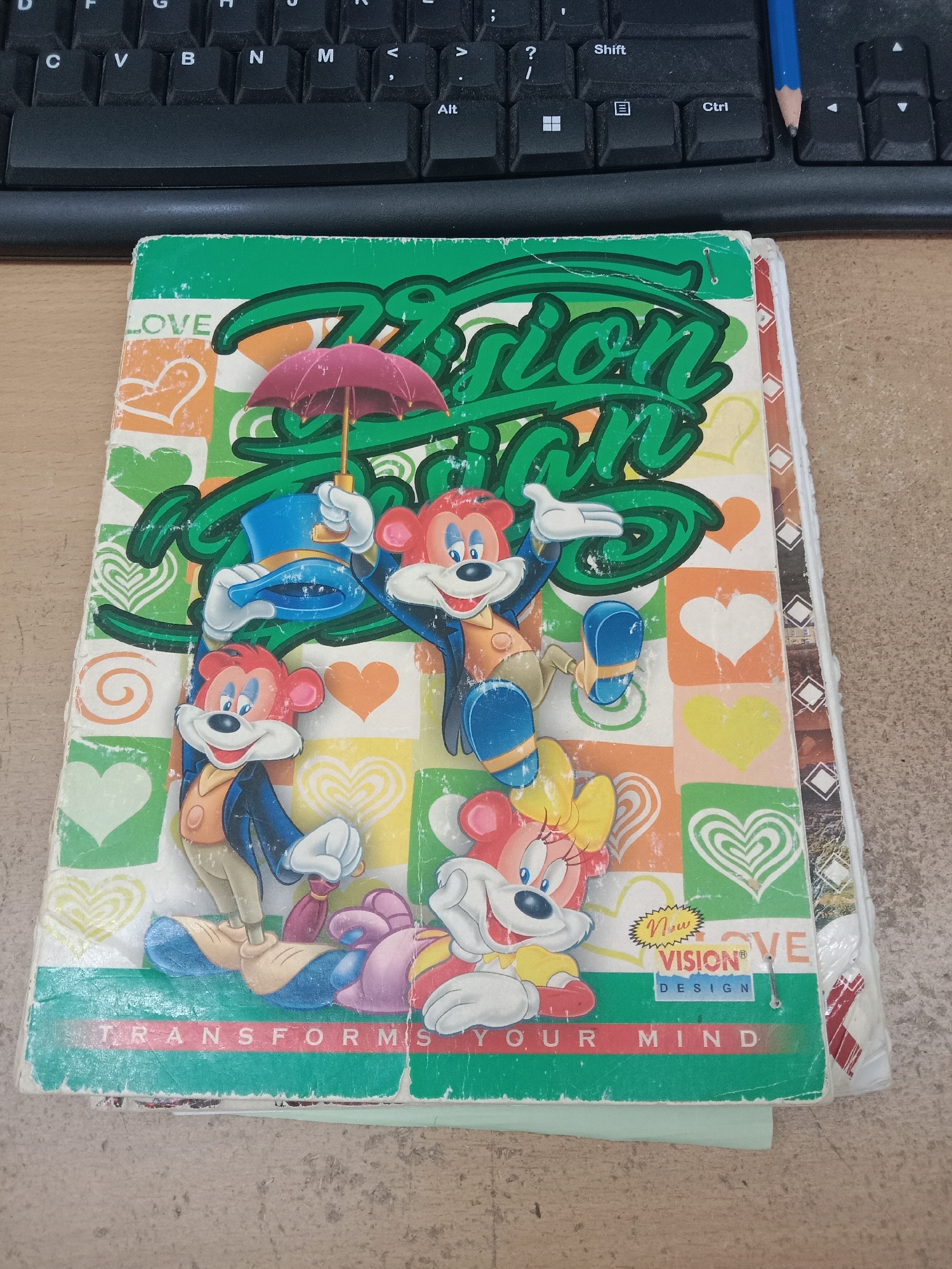 The image shows a closed notebook with a colorful cover, featuring cartoon characters that resemble mice. The cover is primarily green and features a large green text that reads "VISION". Below this, there are smaller text boxes. One box contains "New VISION® DESIGN" with a yellow zig-zag border, and another box has the text "LOVE" to the left of it. The bottom edge of the notebook has green text that reads "TRANSFORM YOUR MIND". The notebook is placed on a wooden surface, likely a desk, with a black computer keyboard visible in the background. A blue pencil with a sharpened tip rests on the keyboard. The notebook cover exhibits signs of wear and tear, with creases and scuffs visible. The image is captured from a slightly overhead angle. There is no indication of the city of Semarang, Indonesia, in the image.