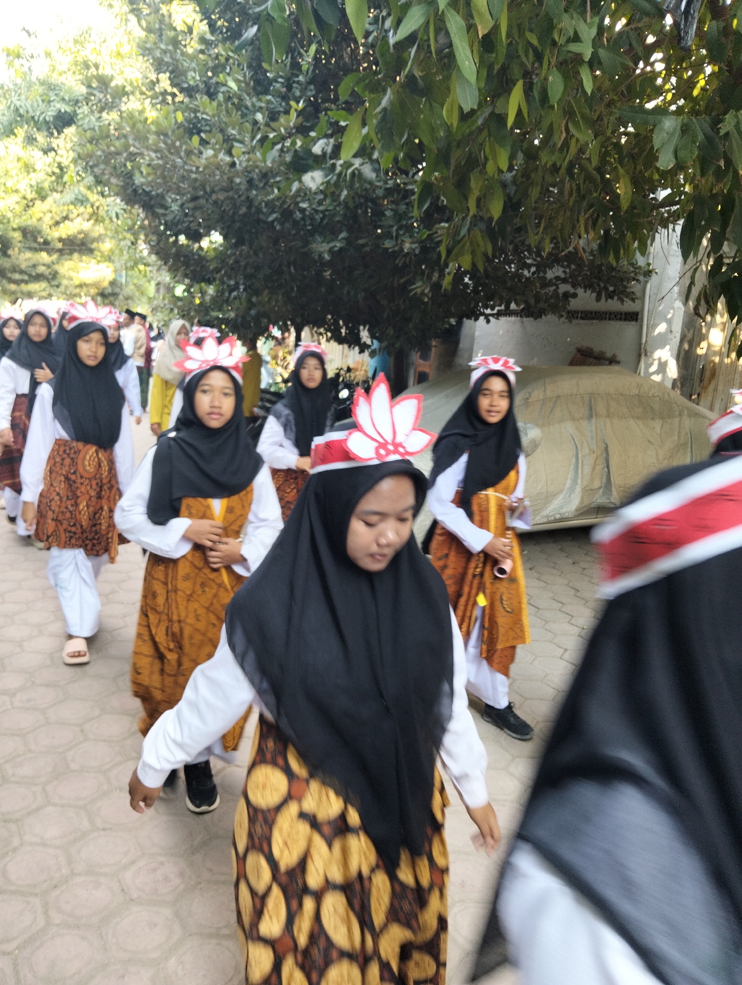 In Kranji, Indonesia, a group of young girls in traditional attire are participating in an outdoor procession. The girls are wearing black hijabs and white shirts, with patterned orange skirts over white pants. Many of them have distinctive flower-shaped headpieces with red and white petals. They are walking on a light-colored, hexagonal-tiled pathway. Trees with lush green foliage line the background, and a light-colored car covered with a tarp is parked to the right. The scene suggests a cultural event or festival taking place in a residential or communal area. The lighting indicates it is daytime, possibly late afternoon due to the soft sunlight filtering through the trees.