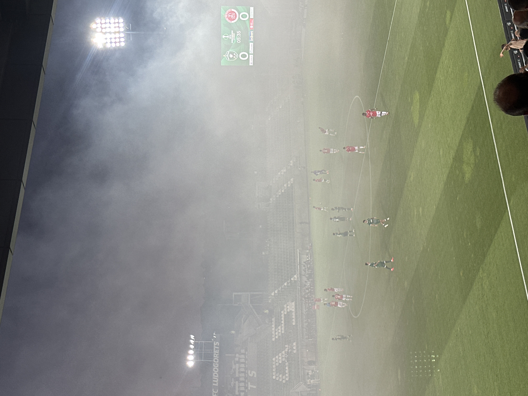 The image depicts a football match taking place at the Huvepharma Arena in Razgrad, Bulgaria. The stadium is characterized by its green football pitch, tiered seating areas, and prominent floodlights. A dense fog or smoke blankets a significant portion of the field and stadium, obscuring visibility. Several players, wearing red and green uniforms, are scattered across the pitch. Some are in motion, suggesting active play, while others appear to be standing or walking. Spectators are visible in the stands, though their view is also impacted by the fog. A large digital scoreboard displays game information, including team scores and the current time, which reads "05:35." The overall atmosphere is hazy and somewhat ethereal due to the atmospheric conditions.