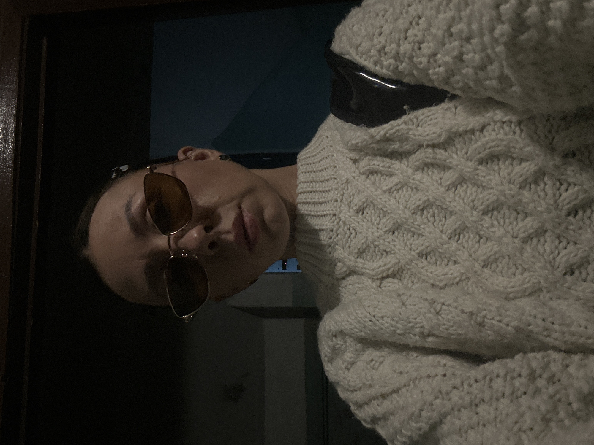 A person wearing sunglasses and a white cable-knit sweater is positioned at a slightly tilted angle in front of a dark doorway. The doorway consists of a dark-colored door and a glimpse of a dark blue wall behind it. The person's face is visible, including their lips, nose, and sunglasses. Part of what appears to be a black bag strap is visible near the shoulder of the sweater. The location is unknown, and there is no information in the image to suggest that the location is in the city of Votkinsk, Russia.