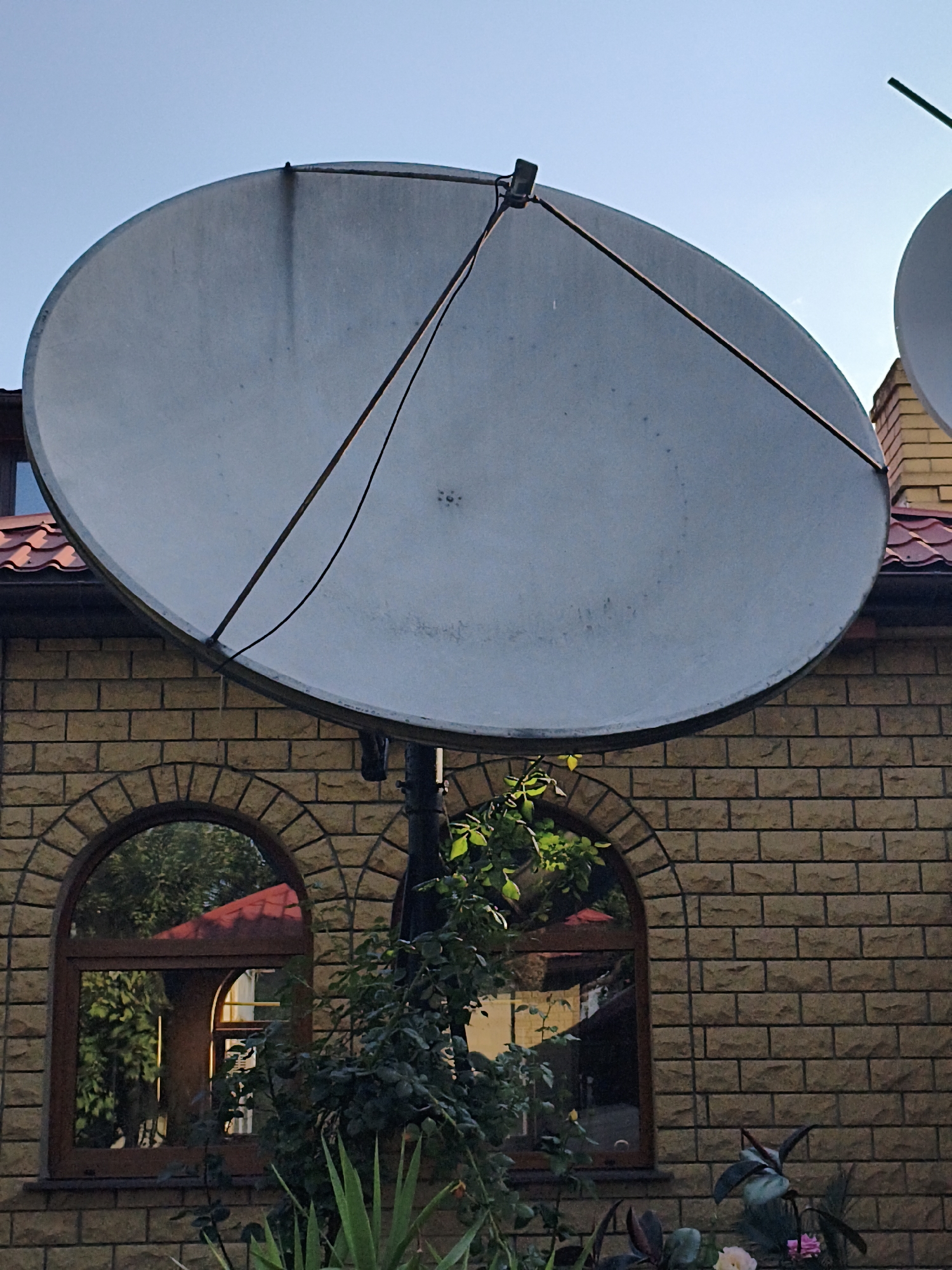 An outdoor, medium shot captures a large, pale satellite dish mounted on a pole, angled upwards against a clear blue sky. The dish occupies the majority of the frame, obscuring the upper portion of a tan brick building with a red tiled roof. The building features two arched windows with reflective glass, showing a distorted reflection of trees and a red roof. Lush green foliage, including plants with spiky leaves and darker, leafy branches, is visible in the foreground, partially obscuring the lower part of the dish and the windows. The overall scene appears to be residential. The location is Makiivka, Ukraine.