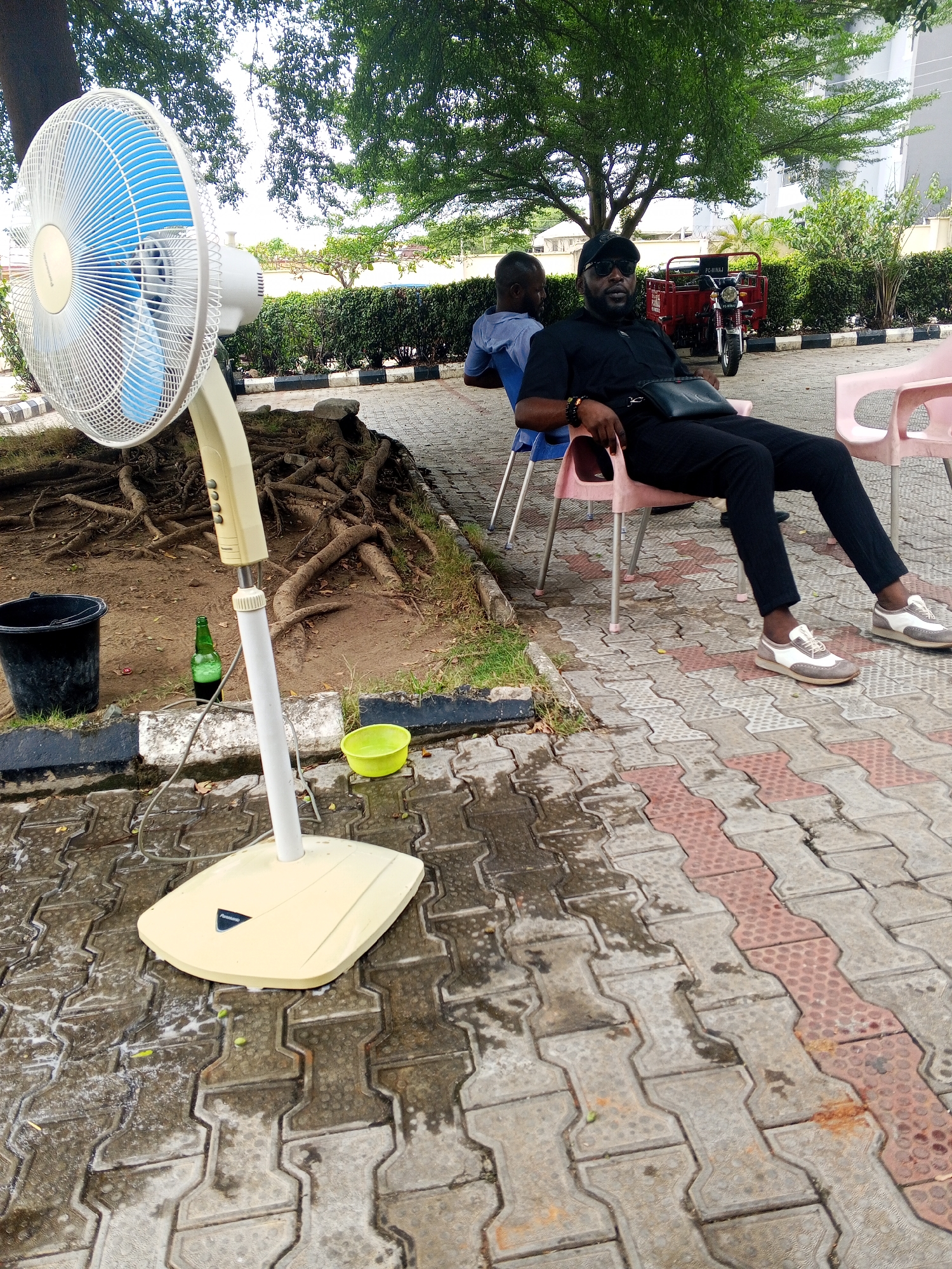 The image shows an outdoor scene in Lokoja, Nigeria. A Black man is seated on a pink plastic chair, wearing all black clothing and sunglasses. Another man, partially visible, is seated on a blue plastic chair behind him. A red three-wheeled vehicle is parked in the background.
In the foreground, there is a standing electric fan with blue blades on a beige base. Adjacent to the fan are a black bucket, a green beer bottle, and a small yellow plastic bowl. The ground is paved with interlocking bricks. The area is bordered by tree roots and greenery.