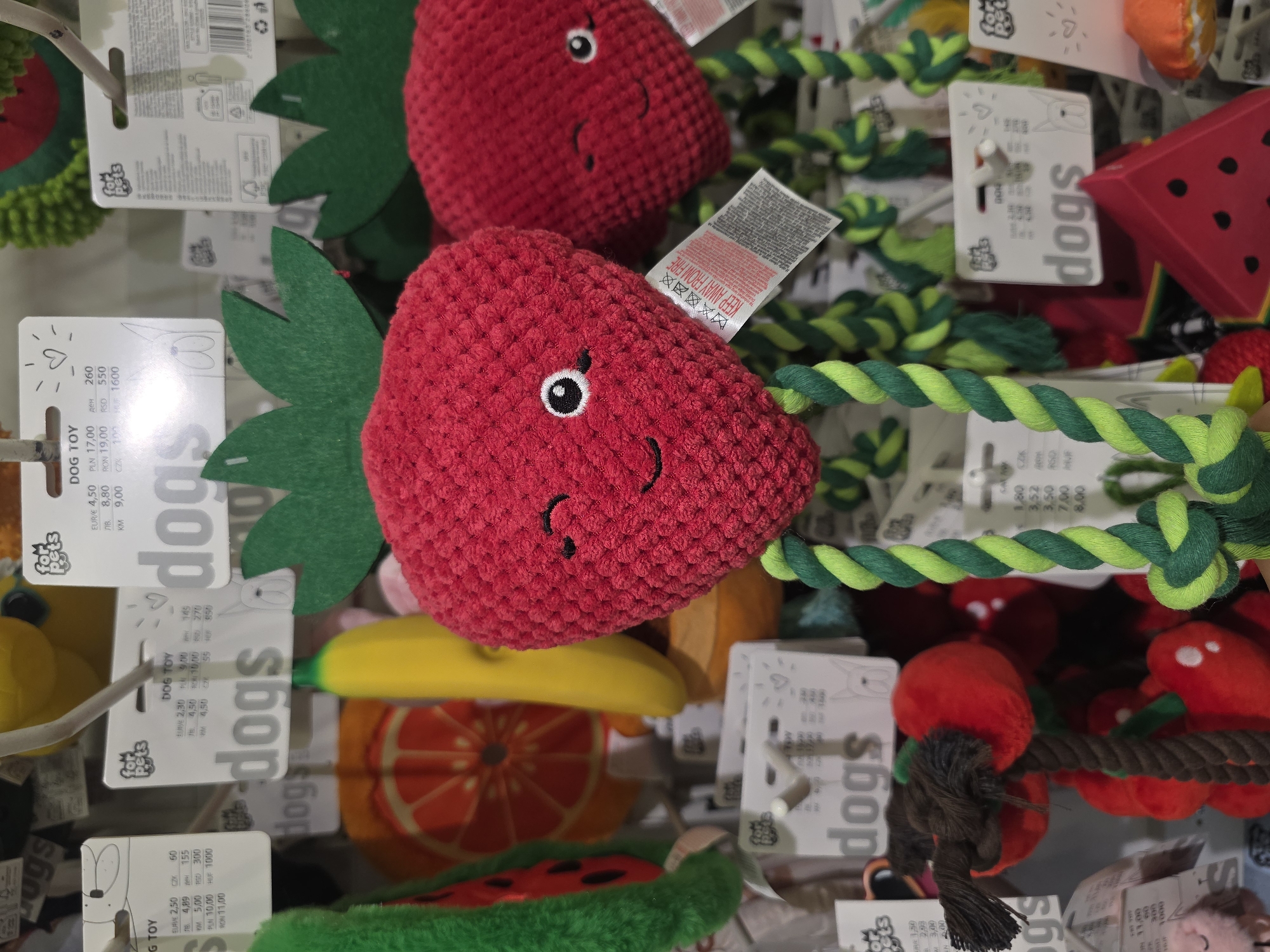 The image shows a display of dog toys, predominantly red strawberry-shaped plush toys with green leafy tops and green and white twisted rope attachments. The toys have embroidered facial features. Many of the toys have "for pets" tags displaying prices in various currencies. Other toys are visible in the background, including a watermelon-shaped toy, a banana, and an orange slice. The presence of Polish currency (PLN) on the tags indicates that the scene is likely in Warsaw, Poland.