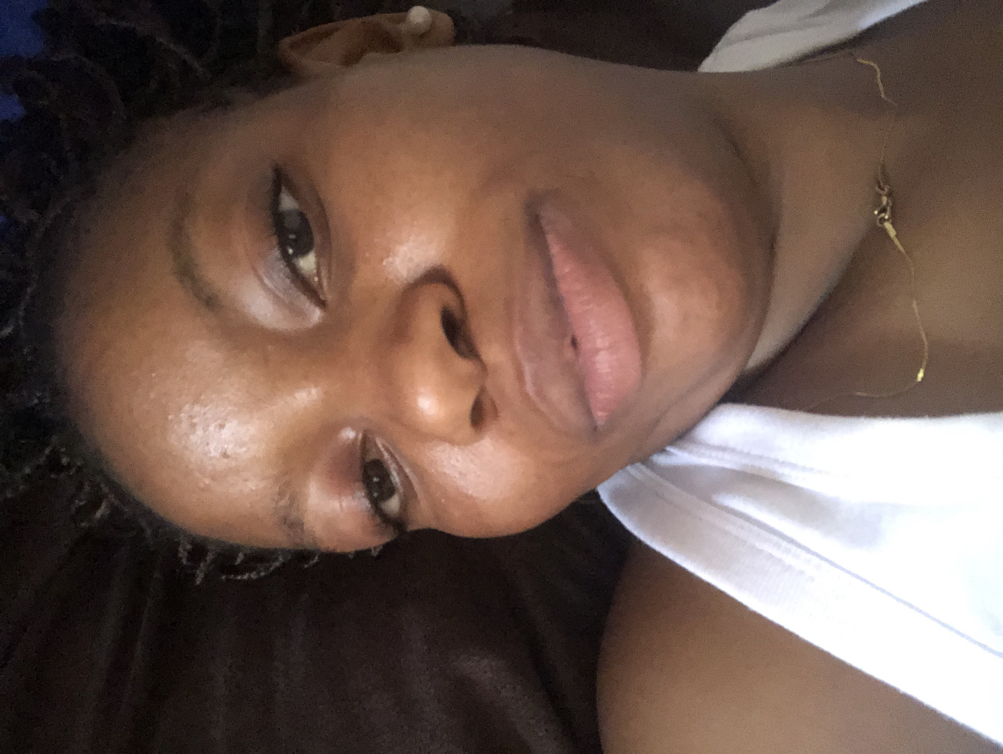 A close-up, angled selfie of a dark-skinned person with short, dark, textured hair, lying on a brown surface, possibly a couch or bed. They are wearing a white tank top and a thin gold necklace. The person's eyes are open, and they are looking slightly downward. The location is unspecified; Kashere, Nigeria, cannot be determined from the image.