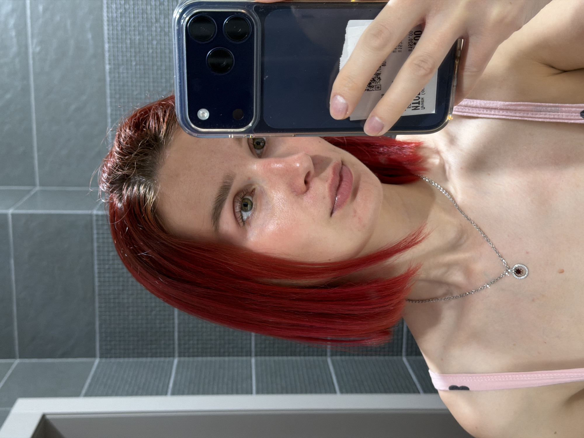 A person with dyed red hair is taking a selfie in what appears to be a bathroom with gray tiled walls. The person is holding a smartphone in a clear case with a dark back, obscuring part of their face. The visible skin appears fair. They are wearing a thin-strapped pink top and a silver necklace with a circular pendant. The person's location or if they are in Chaikovsky, Russia cannot be determined from this image.