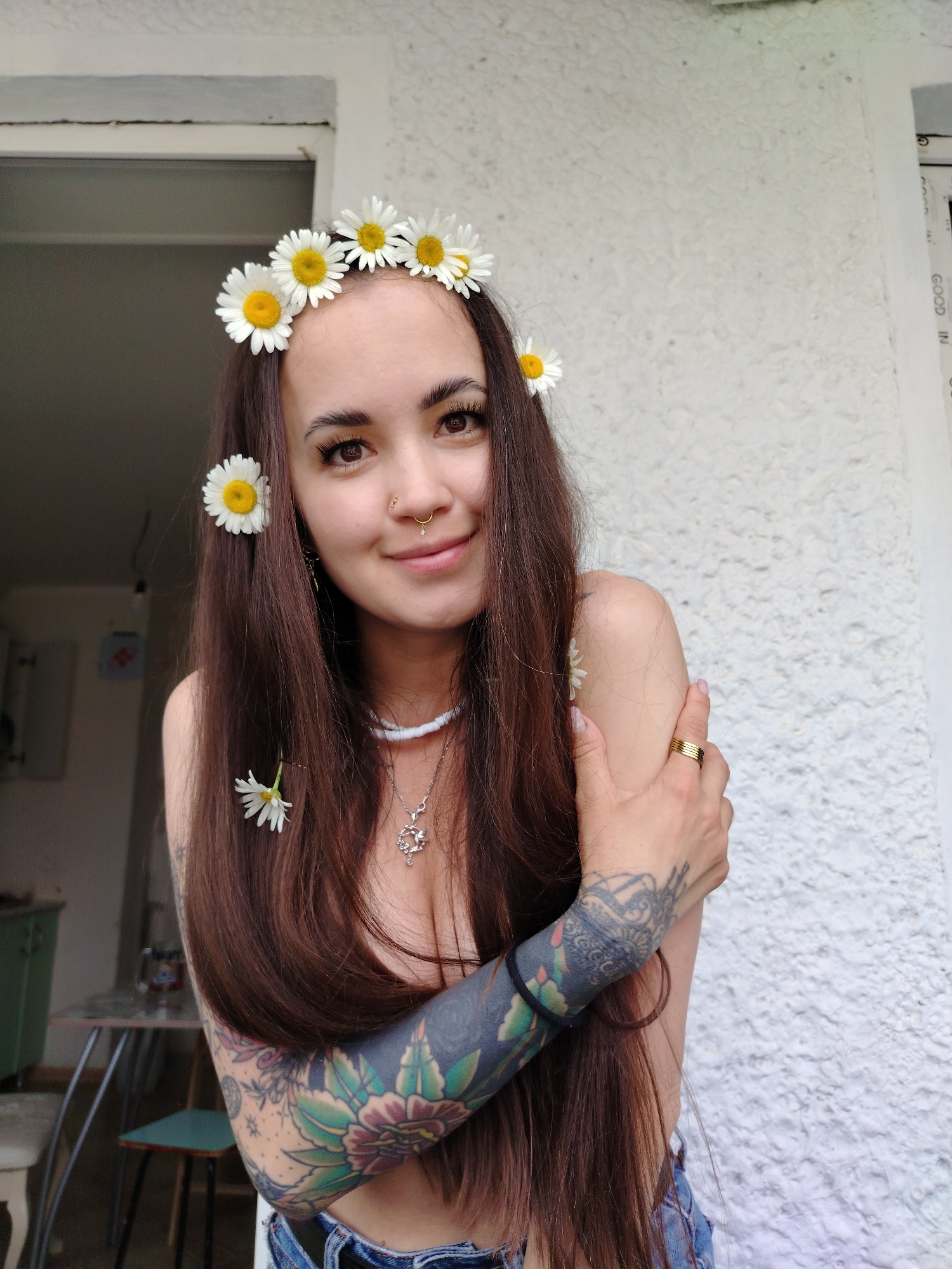 A young woman with long, dark brown hair adorned with a daisy flower crown and a single daisy tucked behind her ear smiles at the camera. She wears a white beaded necklace with a silver pendant, denim shorts, and a gold ring on her right hand. Her left arm and shoulder are covered in colorful tattoos, and her right arm is held across her chest. She is positioned outdoors, with a textured white wall and a window frame visible in the background. The scene suggests a casual outdoor setting, possibly a backyard or patio area in Noviy, Russia, given the context.