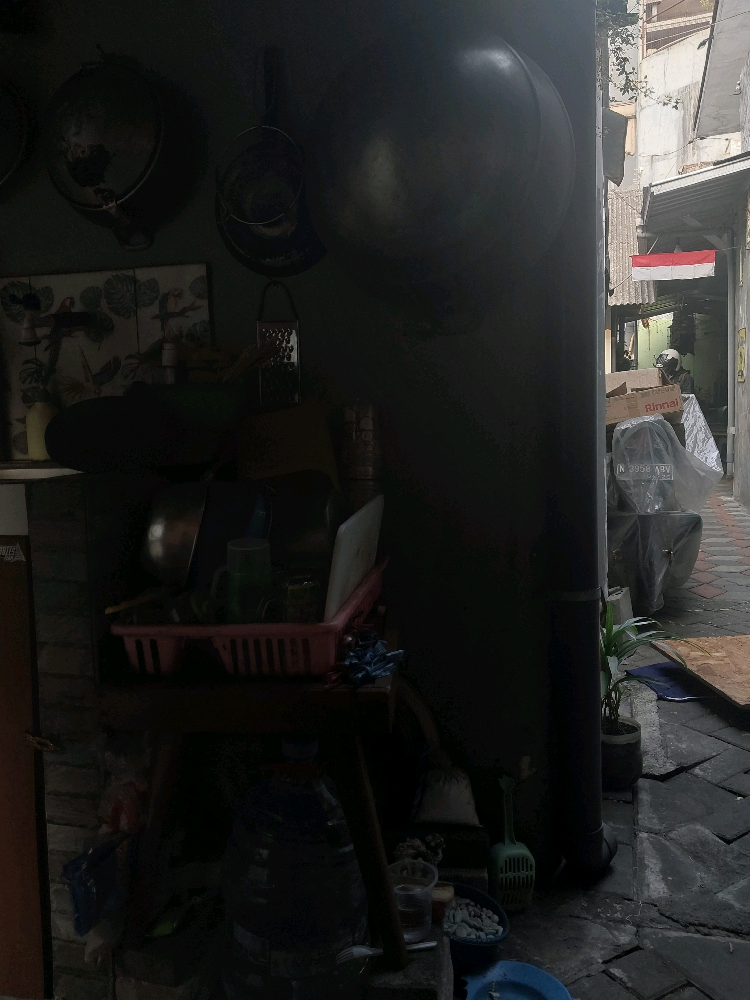 The image depicts an alleyway in a city, likely Malang, Indonesia, based on the presence of an Indonesian flag visible on a clothesline. The scene is dimly lit, with a dark wall dominating the left side of the frame. Various kitchenware, including pots, pans, and bowls, are visible on a shelf. A red plastic dish rack holds cups and other items. In the background, a narrow alleyway opens up, revealing buildings and a paved path. A motorcycle, partially covered with a plastic sheet, is parked on the right side. A person wearing a helmet is seated on the motorcycle. The overall impression is one of a domestic or semi-commercial outdoor space, possibly a kitchen or storage area attached to a residence or shop. The Indonesian flag suggests a cultural context of patriotism or celebration. The scene is a snapshot of everyday life in an urban environment, capturing a moment of stillness and potential activity within a bustling city.
