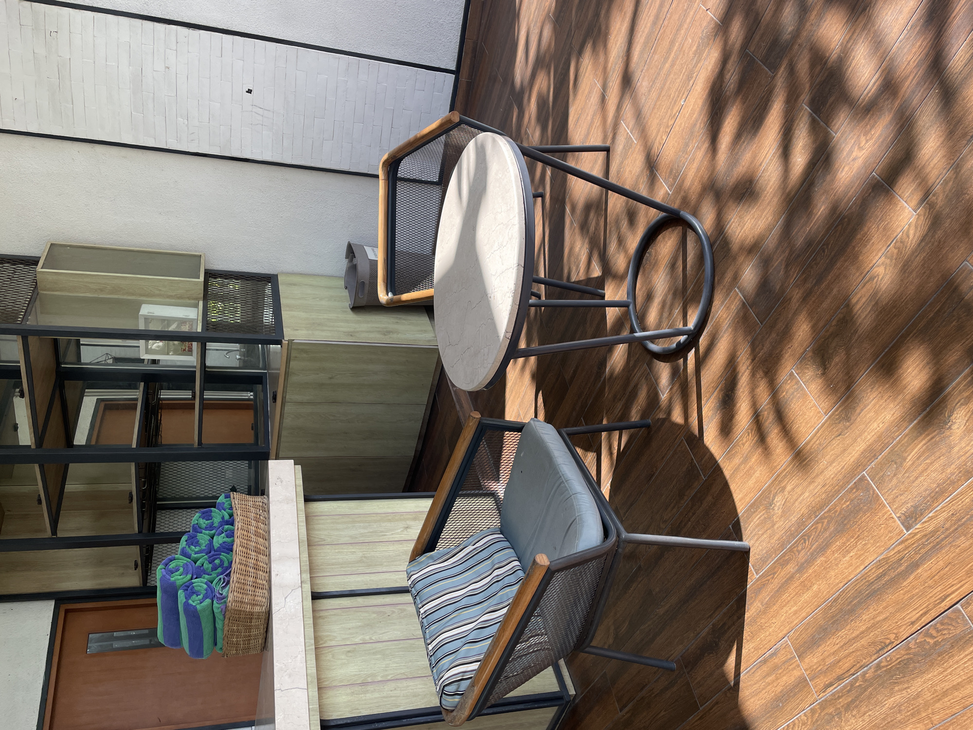 The image shows an outdoor patio area in an unknown city. The flooring is brown wood-look tile laid in a herringbone pattern. A bistro table with a round, light-colored top and dark metal frame stands between two chairs of similar design, featuring mesh backs and cushions. One chair has a blue and white striped pillow. A shadow pattern from a tree or overhead structure is cast across the scene. Against a white brick wall is a multi-tiered shelving unit containing various items, including a basket filled with rolled blue and green towels. A partially visible wooden door is located at the bottom left of the image.