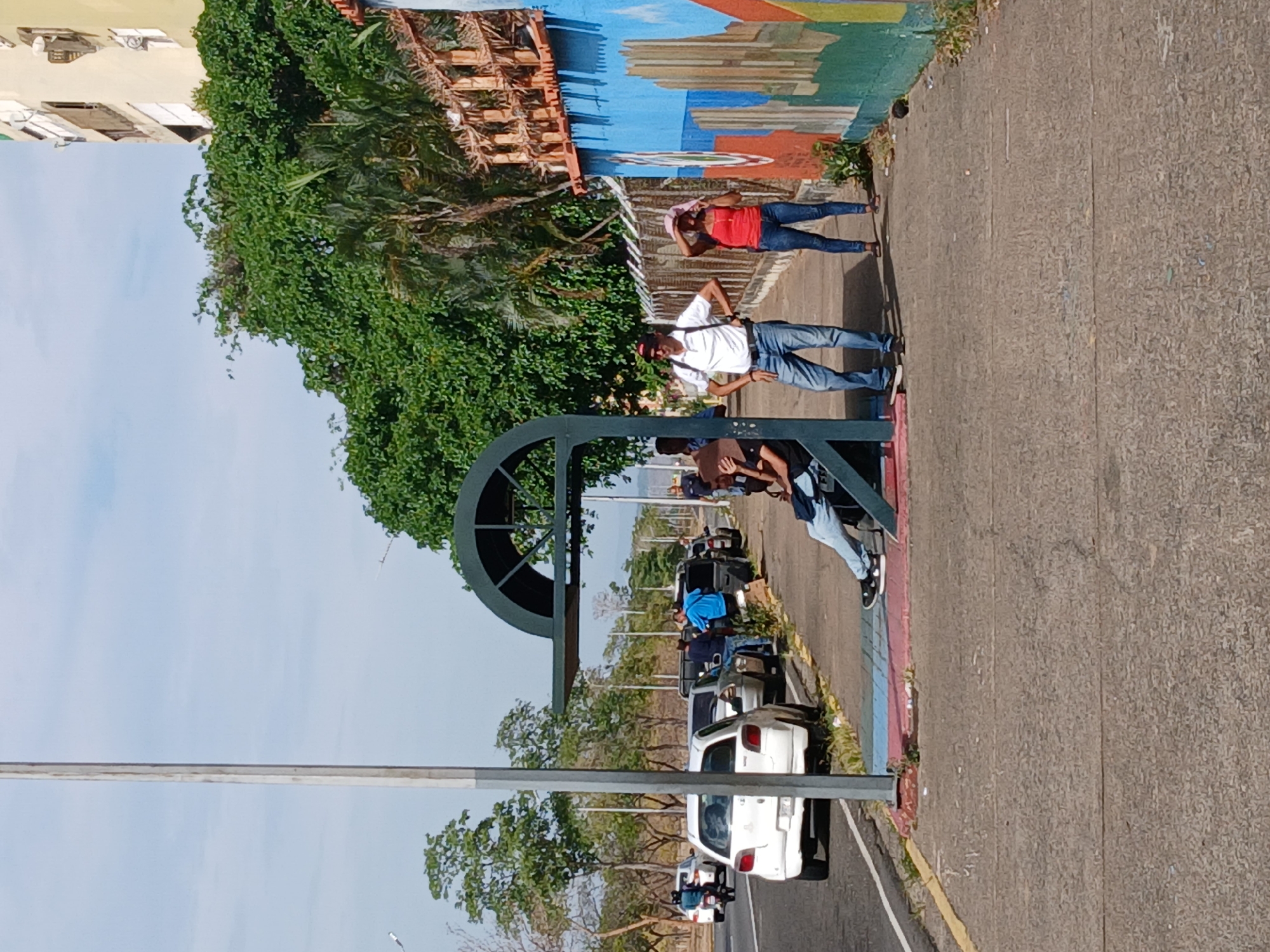 The image captures a street scene in Ciudad Guayana, Venezuela. A group of people are standing on the sidewalk next to a colorful mural. The mural depicts a building under construction, a possible homage to the city's modern architecture. A woman in a red shirt and a pink hat stands near the edge of the sidewalk. A man in a white shirt and blue jeans stands next to her. In the foreground, people are sitting at a bus stop. The street is lined with parked cars, including a white car in the foreground. Trees and greenery are visible in the background, along with a partial view of a building in the distance. The overall environment suggests a typical urban setting.