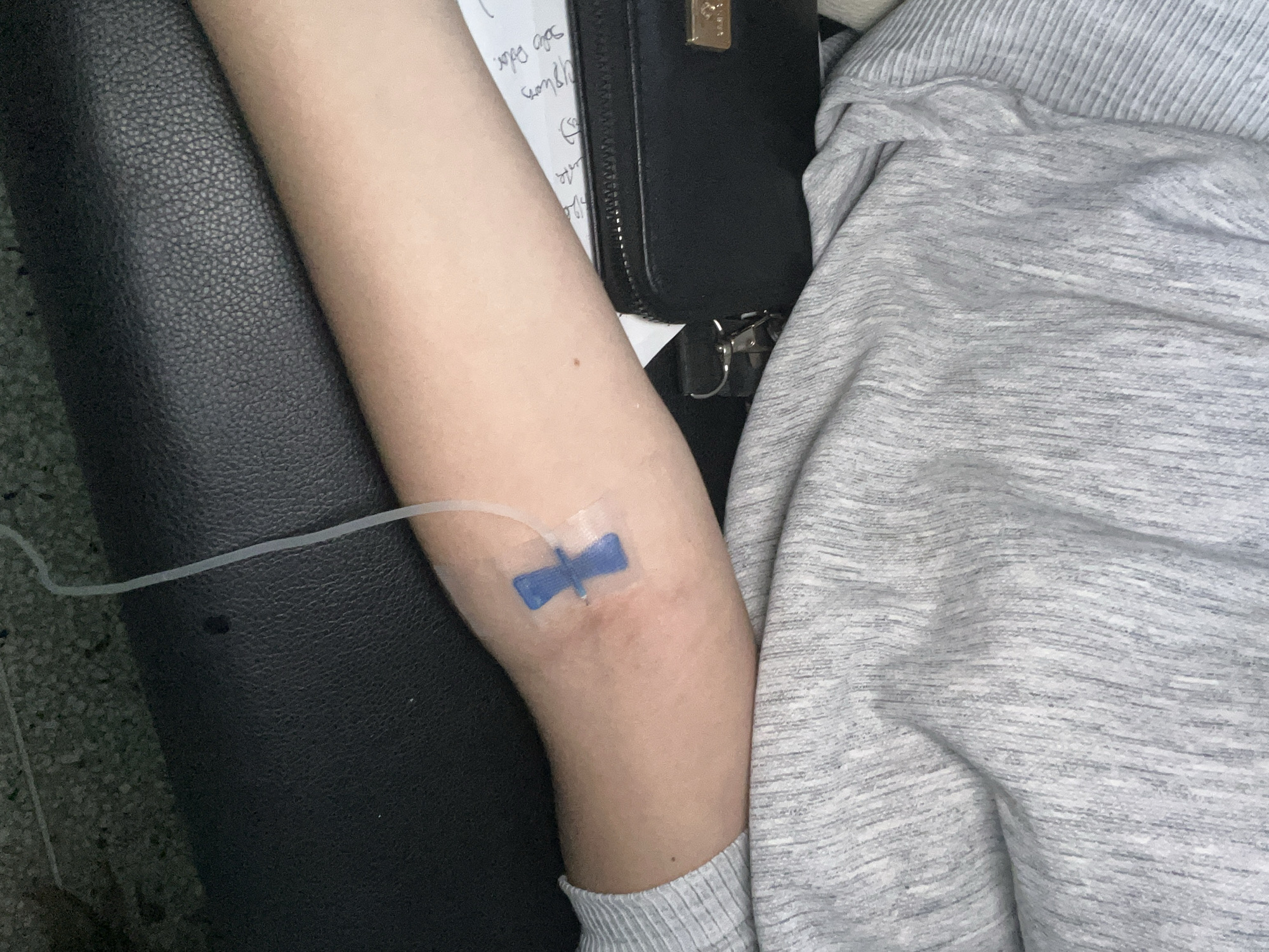 An arm with an intravenous catheter inserted into the inner elbow rests on a black surface, partially covered by a heather gray garment. Next to the arm is a black wallet with a gold clasp and handwritten notes. The location in the city of Juan Griego, Venezuela, cannot be determined from the image.