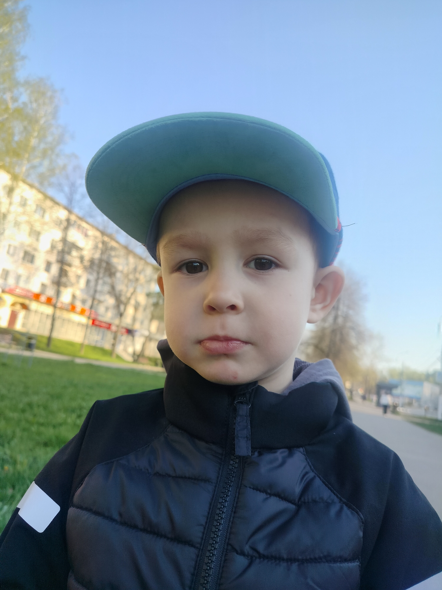 A close-up photograph shows a young child wearing a dark blue and black quilted jacket with a grey hood and a green baseball cap tilted back. The child has light skin, brown eyes, and a neutral expression. The background reveals an urban setting. A light-colored building with red signage and numerous windows is visible on the left. Bare trees are scattered throughout a grassy area. A paved walkway extends into the distance on the right, with blurry figures of people walking. The sky is pale blue. The location is Chaikovsky, Russia, as indicated by the urban environment and architectural style.