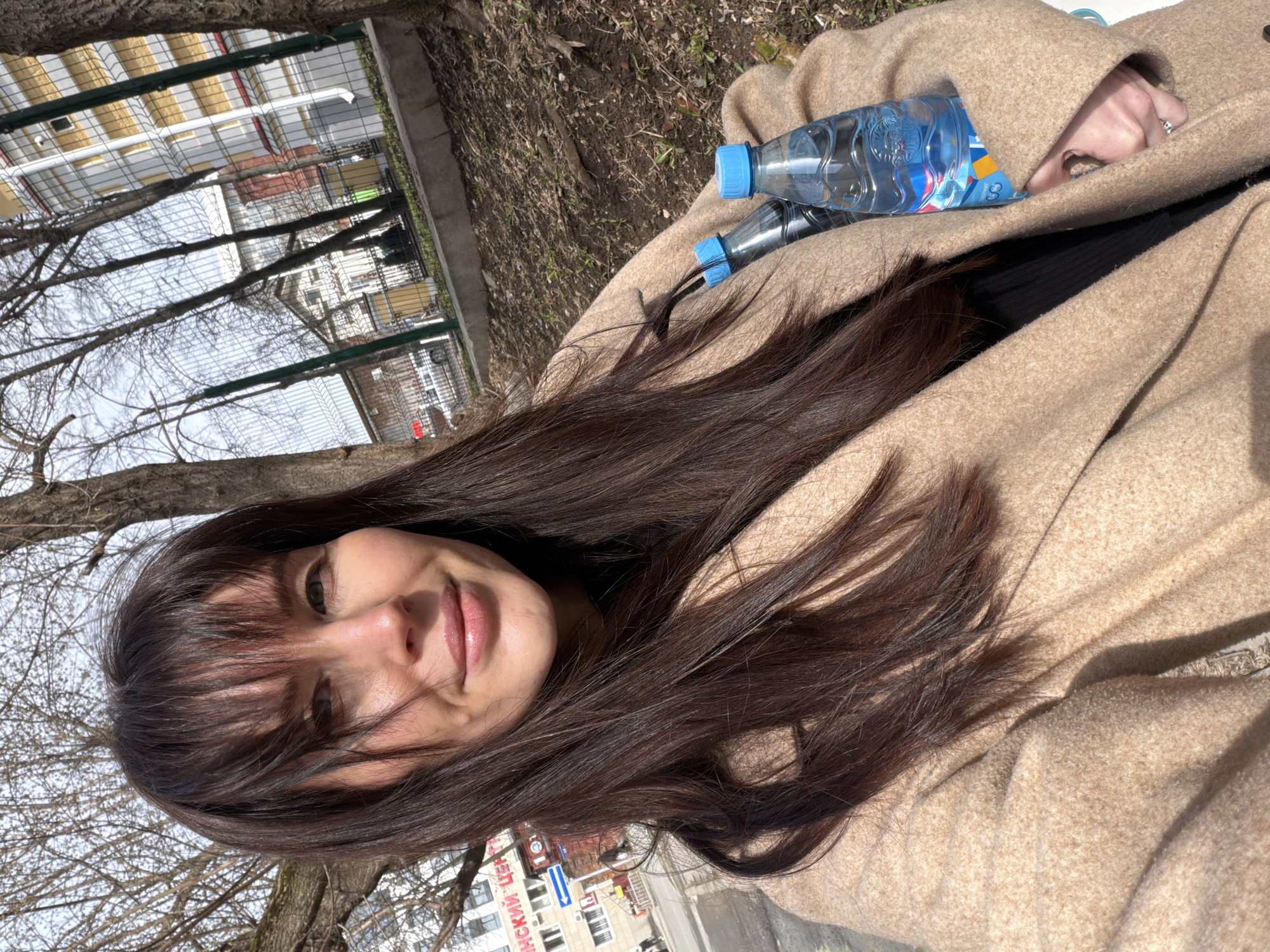 The image is a portrait of a woman taking a selfie or being photographed outdoors on a sunny day in Perm, Russia. The woman, with long, dark brown hair and bangs, is smiling gently at the camera, her face lit by bright sunlight. She is wearing a beige or light brown overcoat, with a dark garment visible beneath.
Two blue-capped plastic water bottles are resting on her coat near her right arm. A silver-colored ring with a stone is visible on a finger of her right hand.
The background shows a city or park setting. Bare tree branches, suggesting late winter or early spring, frame parts of the image. Beyond the trees, there's a clear, light blue sky. In the middle ground, behind a wire mesh fence, buildings and what appears to be a street are visible. One building features a red brick facade. On a building in the distance, a sign with Cyrillic text is partially visible, reading "ИНСКИЙ ЦЕНТР" (referring to a type of "center" in Russian). The ground visible in the background is a mixture of dirt and dry grass. The bright sunlight indicates it is daytime with clear weather.