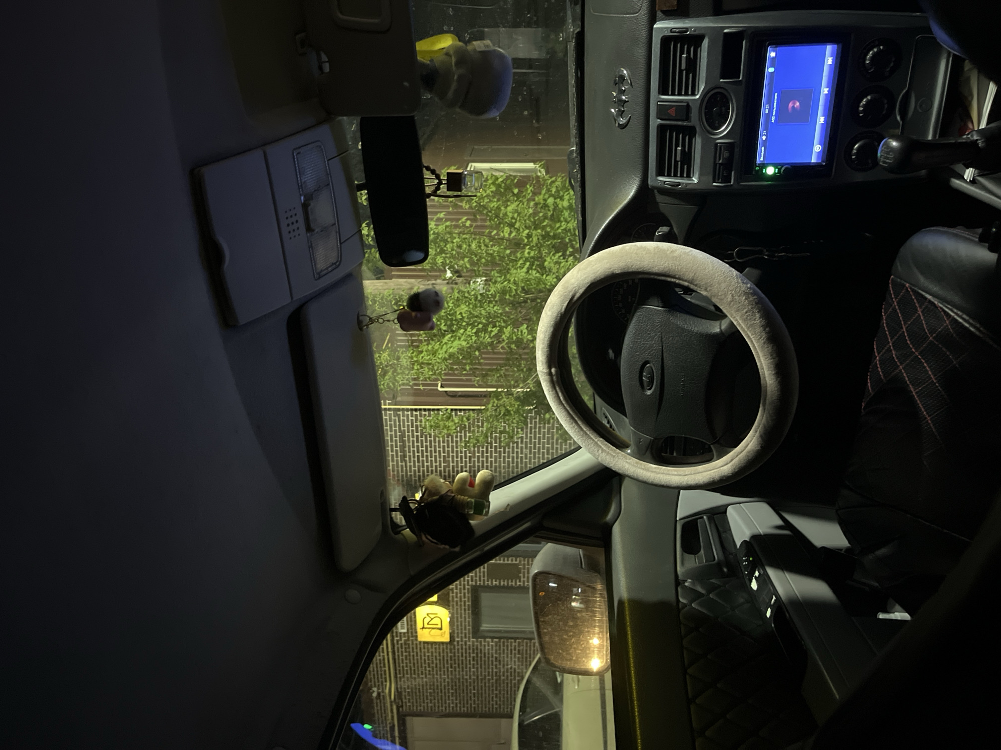 The image depicts the interior of a car, specifically the driver's side. The steering wheel is visible in the center, covered with a light-colored fuzzy material. The dashboard features a digital display screen. The view through the windshield shows a street scene with buildings and greenery. There are also car decorations hanging from the rearview mirror and dashboard. Based on the surrounding environment, the location could be Chaikovsky, Russia.
