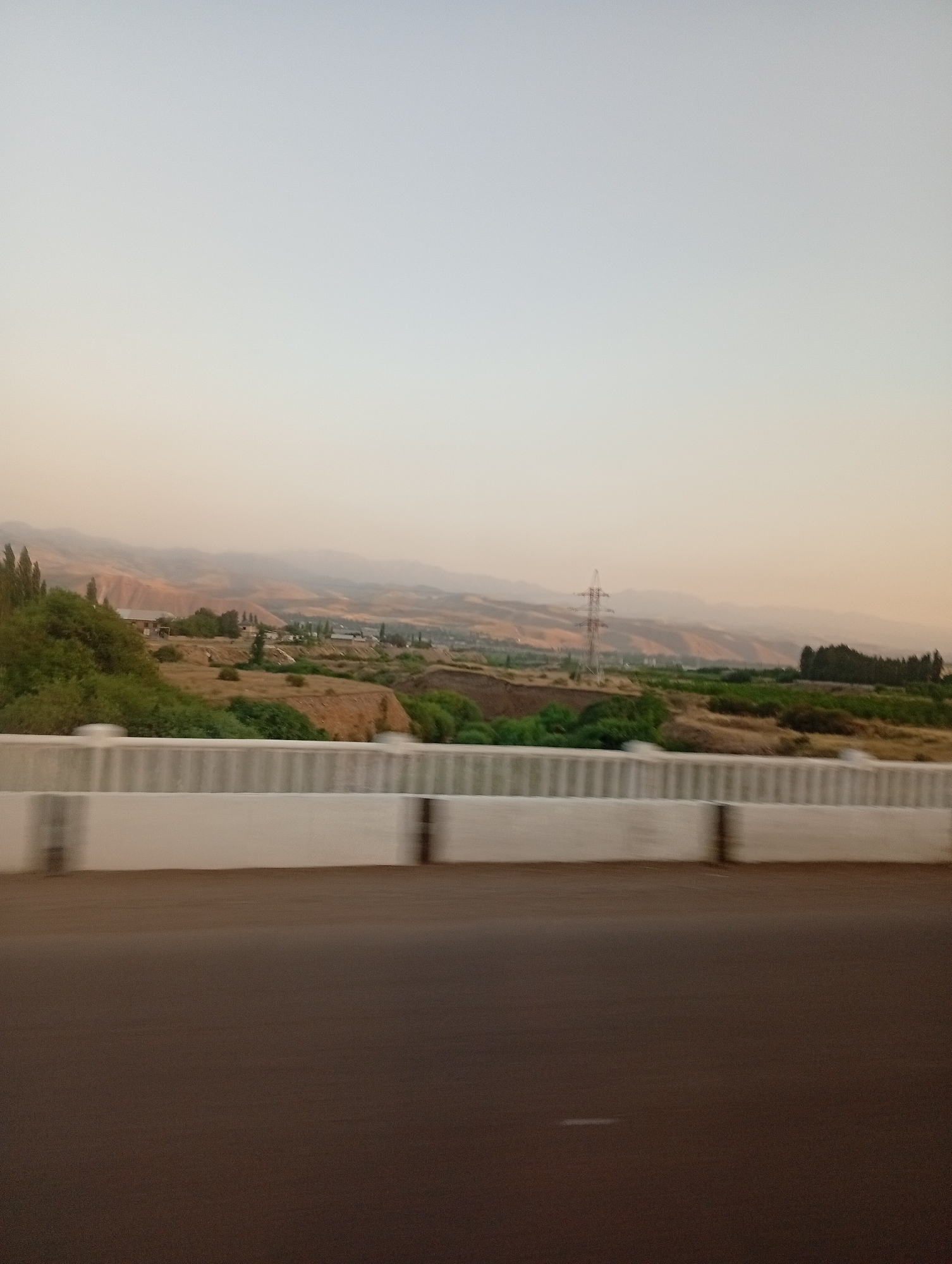 The image depicts a rural landscape with rolling hills and vegetation, likely taken from a moving vehicle. A white concrete barrier with vertical dark stripes runs along the foreground, indicating the viewpoint is from a road or bridge. Beyond the barrier, a dry, grassy hillside is visible, dotted with sparse trees and some low-lying buildings. Further in the distance, hazy mountains form the backdrop under a pale, overcast sky. A single, tall electrical pylon stands prominently on one of the hills. The overall impression is one of a natural, somewhat arid environment, possibly on the outskirts of a settlement in Angren, Uzbekistan.