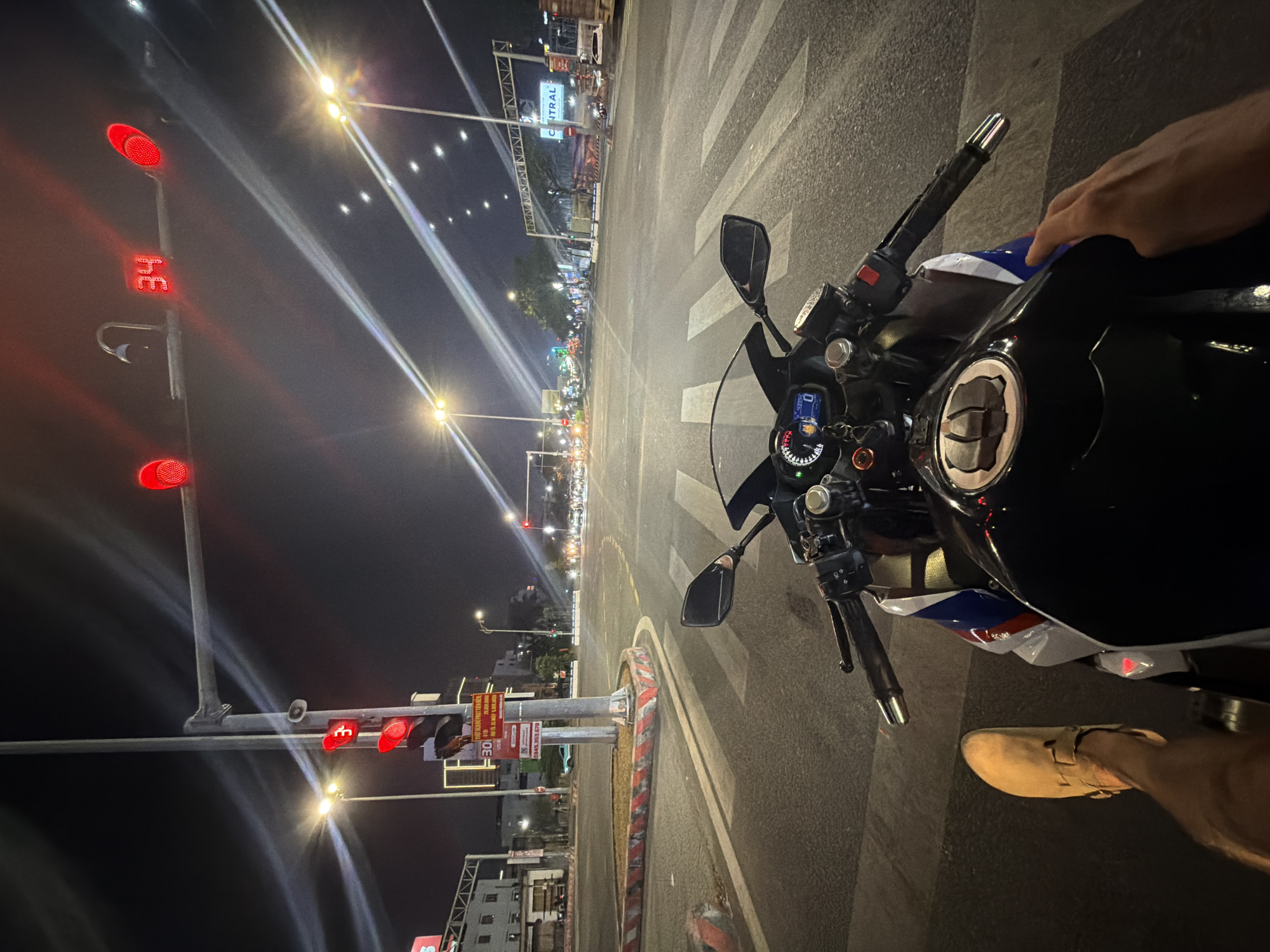 View from the seat of a motorcycle, facing towards the "Central" shopping mall in Đà Nẵng, Vietnam, at night. The motorcycle's handlebars, mirrors, dashboard, fuel tank, and the rider's hands are visible. The motorcycle is stopped at a red traffic light. Traffic lights are visible on the left. The roadway is asphalt. There is pedestrian crosswalk. A sign is visible on the right. Street lights illuminate the scene. A shoe and the rider's leg are visible.