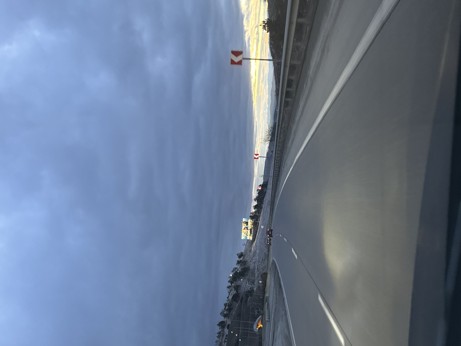 An overcast sky looms over a curved highway. The road surface reflects light, suggesting wet conditions. Signs with red chevrons are posted along the roadside. A billboard is visible in the distance, along with sparse vegetation that appears frosty or snow-covered. The photograph appears to have been taken from inside a vehicle.