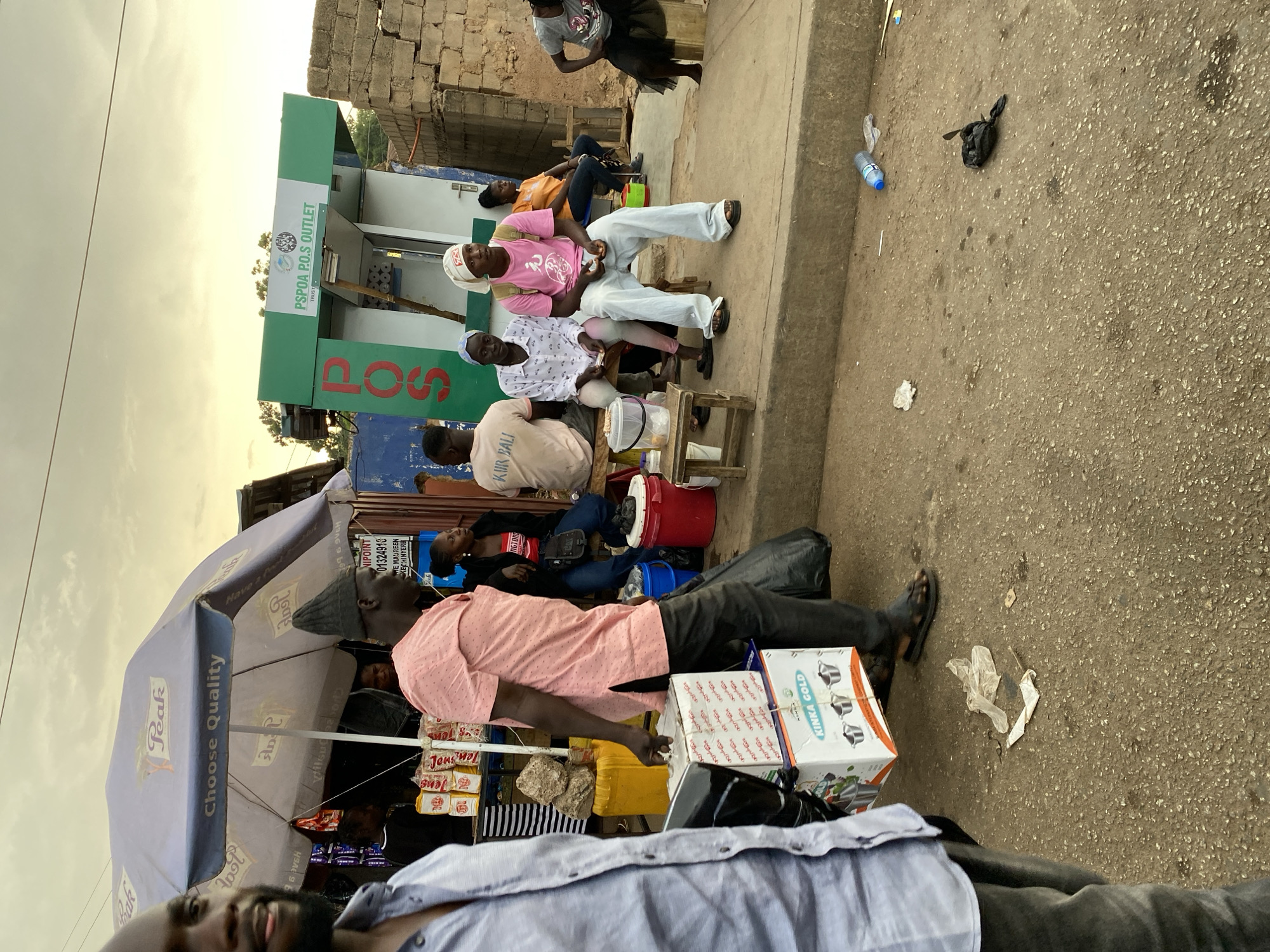 The image captures a street scene in Bukuru, Nigeria, featuring a mix of commercial and social activity. On the left, a "Peak Choose Quality" umbrella shades a small business offering "KINKA GOLD" products and "Jons Jols" items. A man in a pink shirt stands near boxes, while another man in a light blue shirt is partly visible in the foreground. Further back, several individuals are sitting outside a green "PSPOA P.O.S OUTLET TRUST" building. Some appear to be drinking from cups or interacting. The street shows signs of activity, with scattered trash, a water bottle, and a black plastic bag. A few people are standing in the background near a concrete building. The scene suggests a typical day in a busy urban setting.