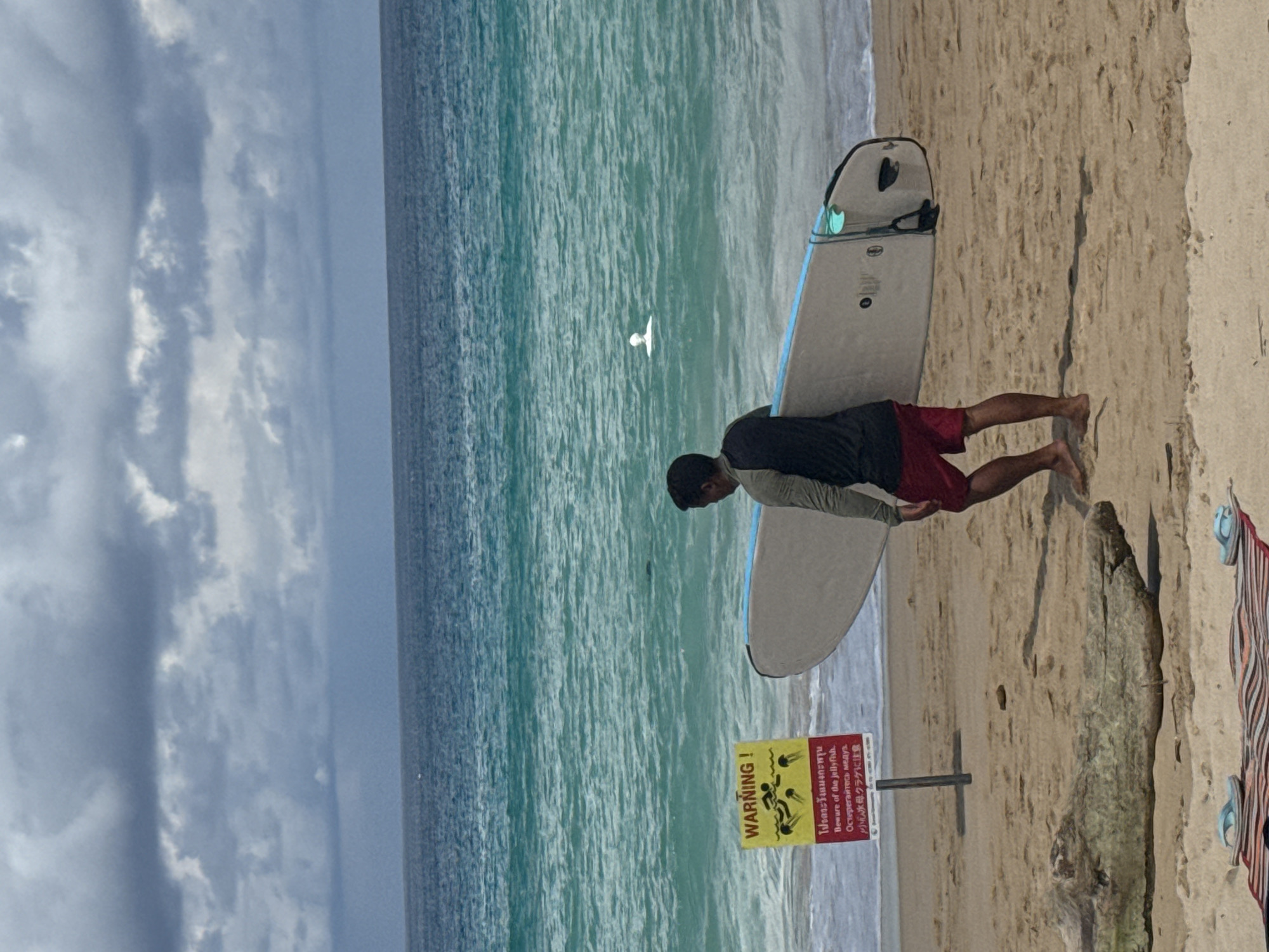 A person walks along a sandy beach carrying a surfboard. The person is wearing a dark long-sleeved shirt and red shorts. The surfboard is light-colored with blue trim. In the background, the ocean is turquoise, and there are dark clouds in the sky. To the left of the person is a yellow warning sign with text and an image of jellyfish. A large piece of driftwood lies on the sand in the foreground. Two pairs of flip-flops are visible on a striped mat near the bottom of the image. The scene appears to be a tropical beach.