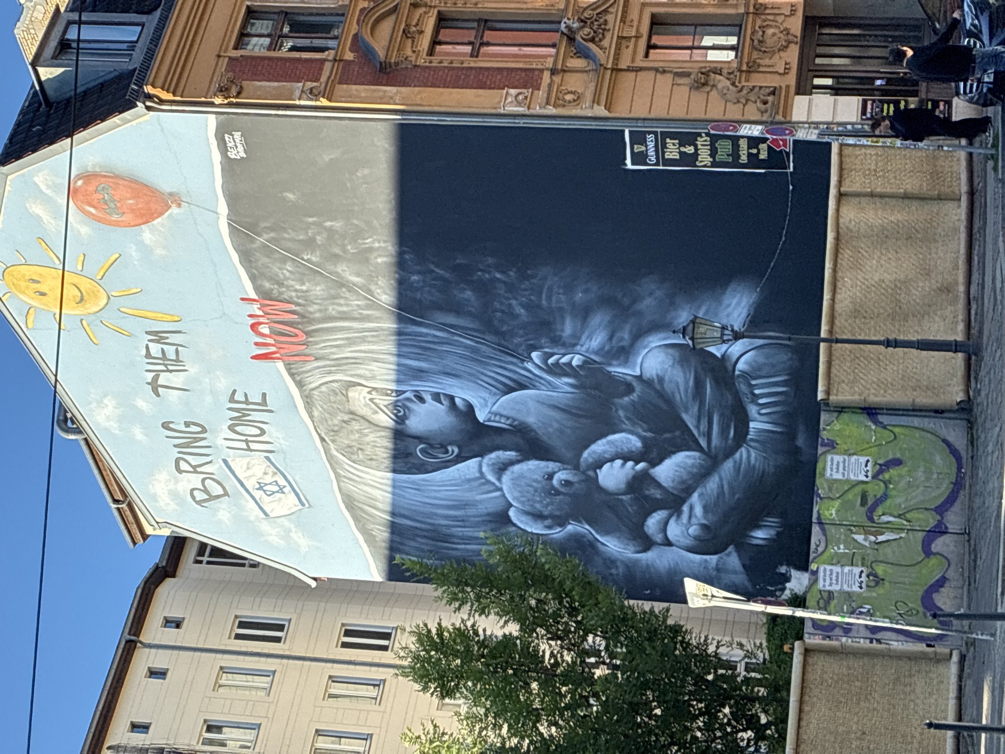 The image shows a mural on the side of a building in Berlin, Germany. The mural depicts a young boy holding a teddy bear, with the words "BRING THEM HOME NOW" written above him. There is also a sun and a red balloon in the sky. The building itself appears to be old, with ornate architecture. In the foreground, there is a street with a lamppost and a few people walking. There is also a sign for a pub that says "Guinness Bier & Sports-Pub". The overall scene is somewhat somber, given the message of the mural.