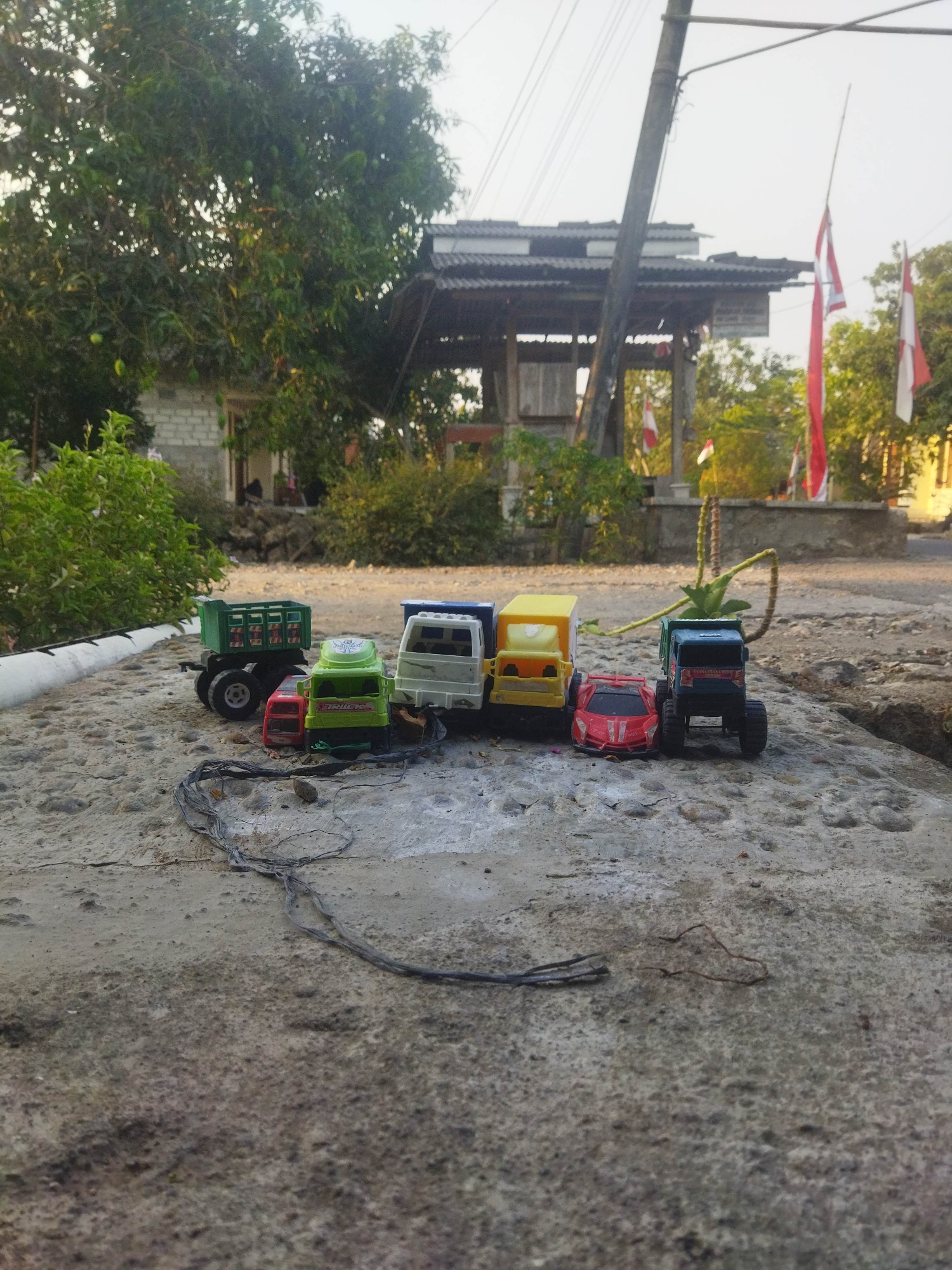 A low-angle shot captures a collection of toy vehicles arranged on a cracked concrete surface, likely in the city of Suci, Indonesia, given the presence of Indonesian flags in the background. The toys include a green dump truck, a red car, a green truck labeled "TRUCK", a white truck, a yellow box truck, and a dark blue truck. Behind the toys, a lush green tree with visible fruit and a building with a corrugated roof and white brick walls are discernible. A prominent wooden pole with electrical wires stretches across the frame, and several red and white Indonesian flags are mounted on poles in the middle ground. The environment appears to be outdoors, with dry ground and vegetation surrounding the scene. The lighting suggests it is daytime, with soft, diffused light. No people are visible in the image.
