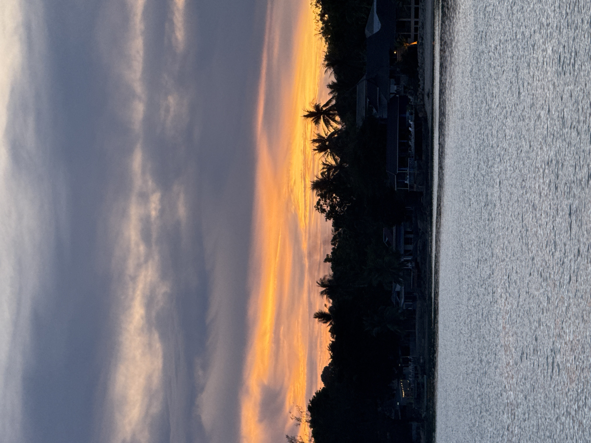 The image depicts a sunset scene over water, likely a bay or calm ocean, in Baan Tai, Thailand. The sky exhibits a dramatic gradient, transitioning from a dark, cloudy blue on the left to a vibrant orange and yellow near the horizon. Silhouetted palm trees and buildings line the shoreline, creating a dark contrast against the colorful sky. The water surface reflects the sky's colors, with subtle ripples adding texture. There are no people visible in the frame.
