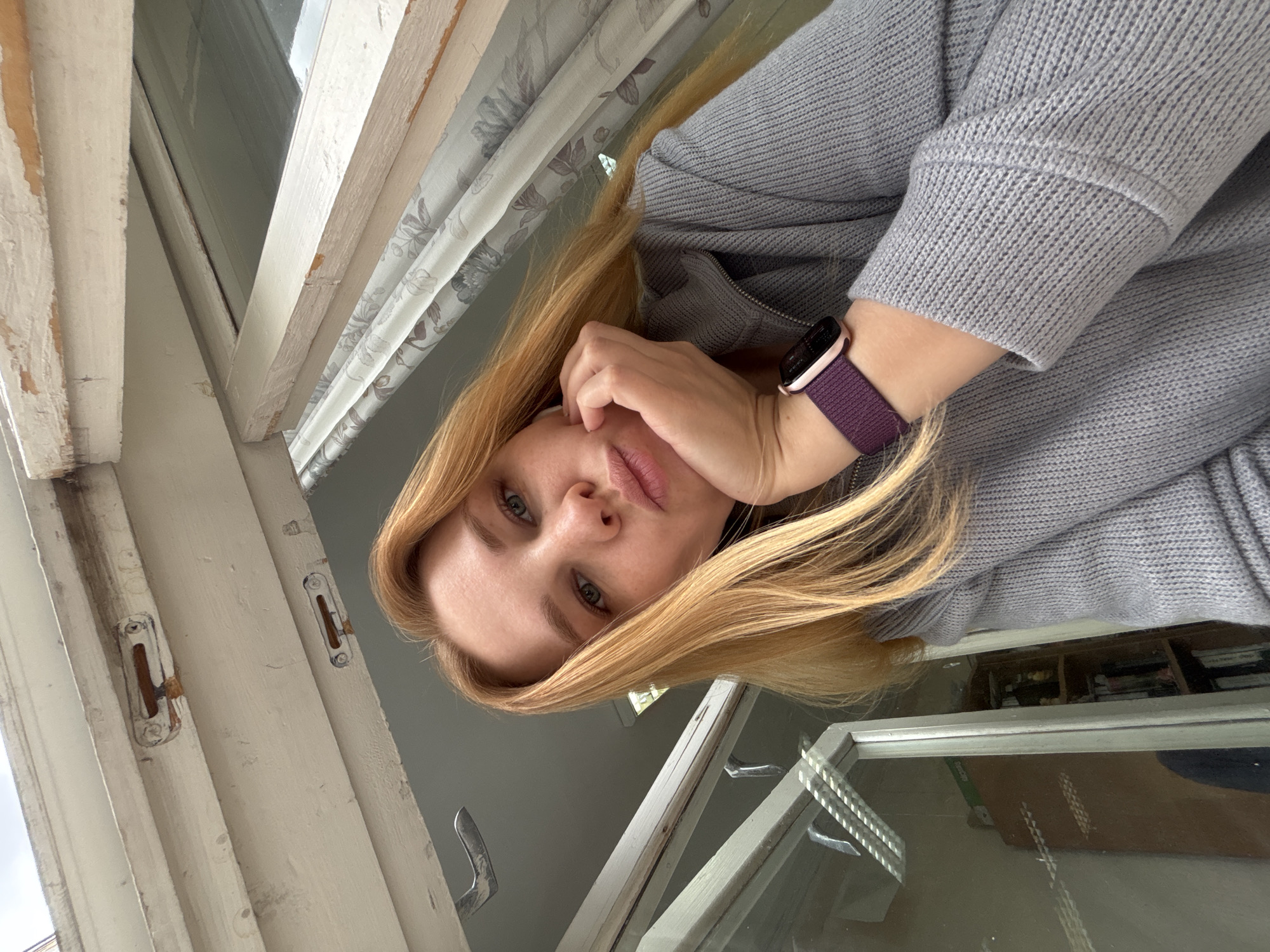 A woman with blonde hair, wearing a grey sweater and a smartwatch with a purple band, poses for a selfie by an old white-framed window. The window frame shows signs of aging, with cracked and peeling paint. A sheer curtain with a floral pattern hangs behind the woman. An interior view of a room is visible through the window. Location cannot be verified in Rusanovo, Russia.