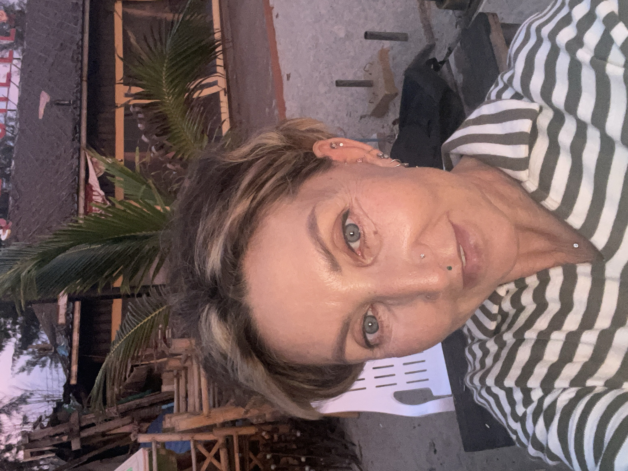A woman with short, blonde-streaked hair is taking a selfie outdoors. She is wearing a green and white striped shirt. She has piercings in her ears and nose. In the background, there is a palm tree, some rustic wooden structures, and a building with a sign that appears to read "HOTEL". The lighting suggests it is either dawn or dusk. The scene is consistent with a beach resort environment in Choeng Thale, Thailand.