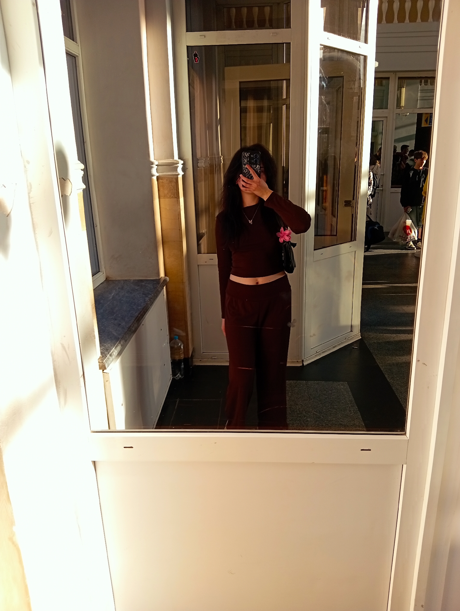 A young woman in a maroon tracksuit stands in front of a mirror, obscuring her face with a smartphone while taking a picture of herself. The reflection reveals an entrance area, possibly a railway station, with other people visible in the background. The location is suggested to be in Poltava, Ukraine, but there is no clear indication of this in the image.