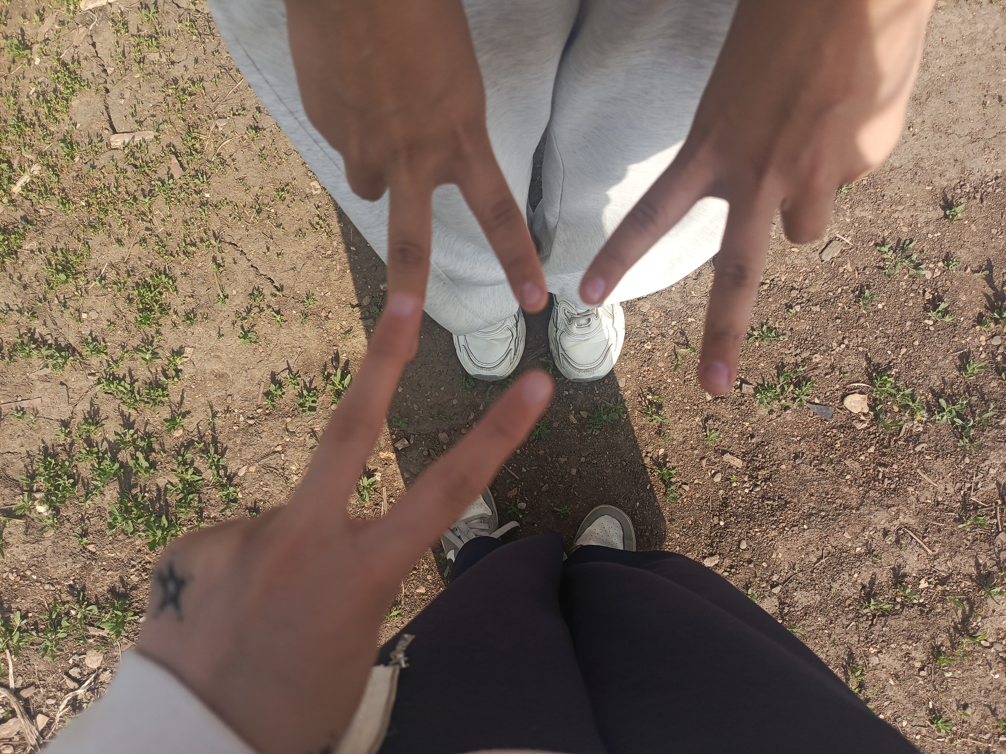 The image shows a view from above of two people standing on ground covered in dry dirt and sparse vegetation. The person in the foreground is wearing dark-colored pants and light-colored sneakers. Their left hand, adorned with a star tattoo on the wrist, is raised to display a "peace" or "victory" sign. The person in the background is wearing light gray sweatpants and white sneakers. Their hands are also raised, making the same "peace" or "victory" sign. The location appears to be outdoors, possibly in a park or unkempt area in an unidentified city, with natural light indicating daytime.