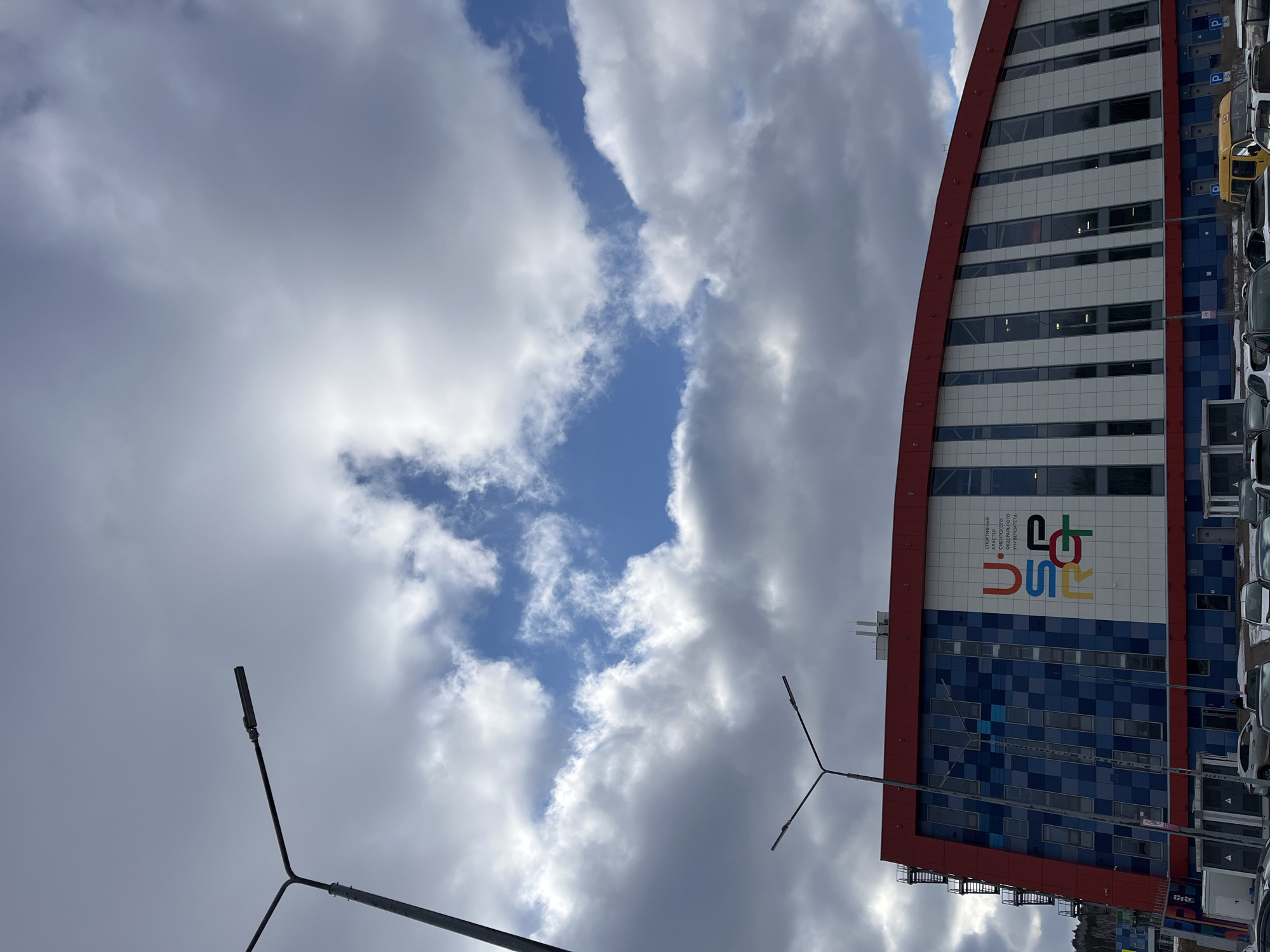 The image depicts the "Cluster Raduga" sports complex in Krasnoyarsk, Russia. The building is multi-storied, with a curved facade featuring a red trim, white and grey tiled sections, and a blue mosaic patterned base. The logo of the Siberian Federal University (SFU) is displayed prominently on the building's facade. To the right of the building, a parking lot with numerous cars is visible. In the foreground, there are two street lights. The sky is cloudy with patches of blue visible in between the clouds. The photo is taken from a low angle, looking up at the building and sky.