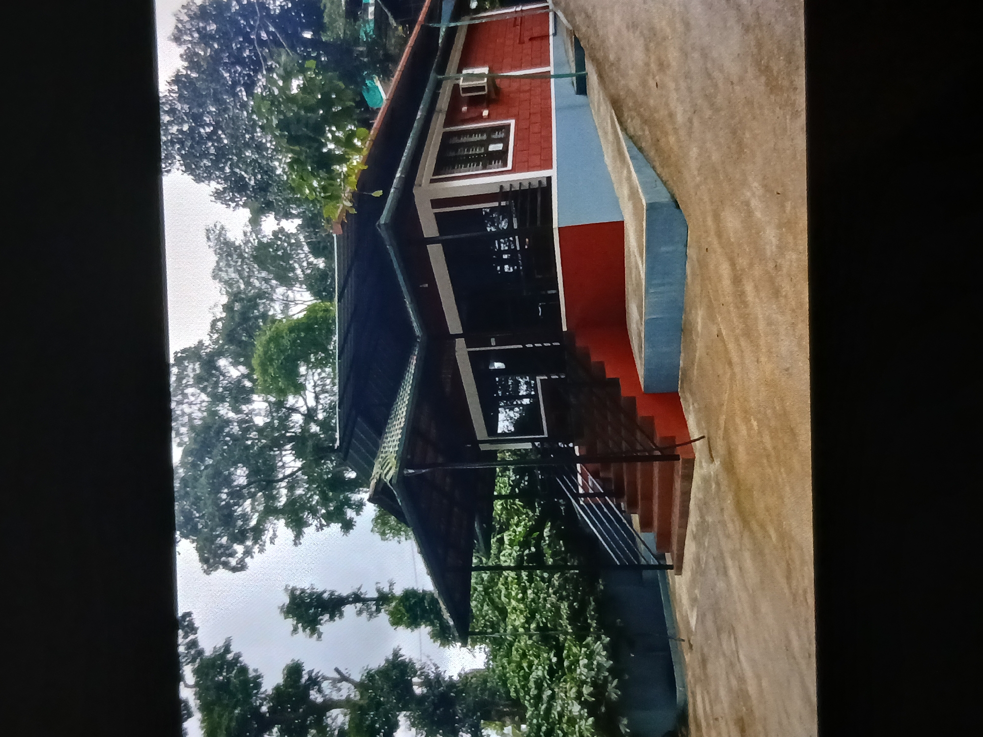 The image shows a two-story house with a red and blue exterior. The house has a balcony with black railings and a staircase leading up to it. There are trees surrounding the house. The location is likely Bengaluru, India, given the information provided, although there is no specific indicator. The house appears to be situated in a somewhat natural environment.