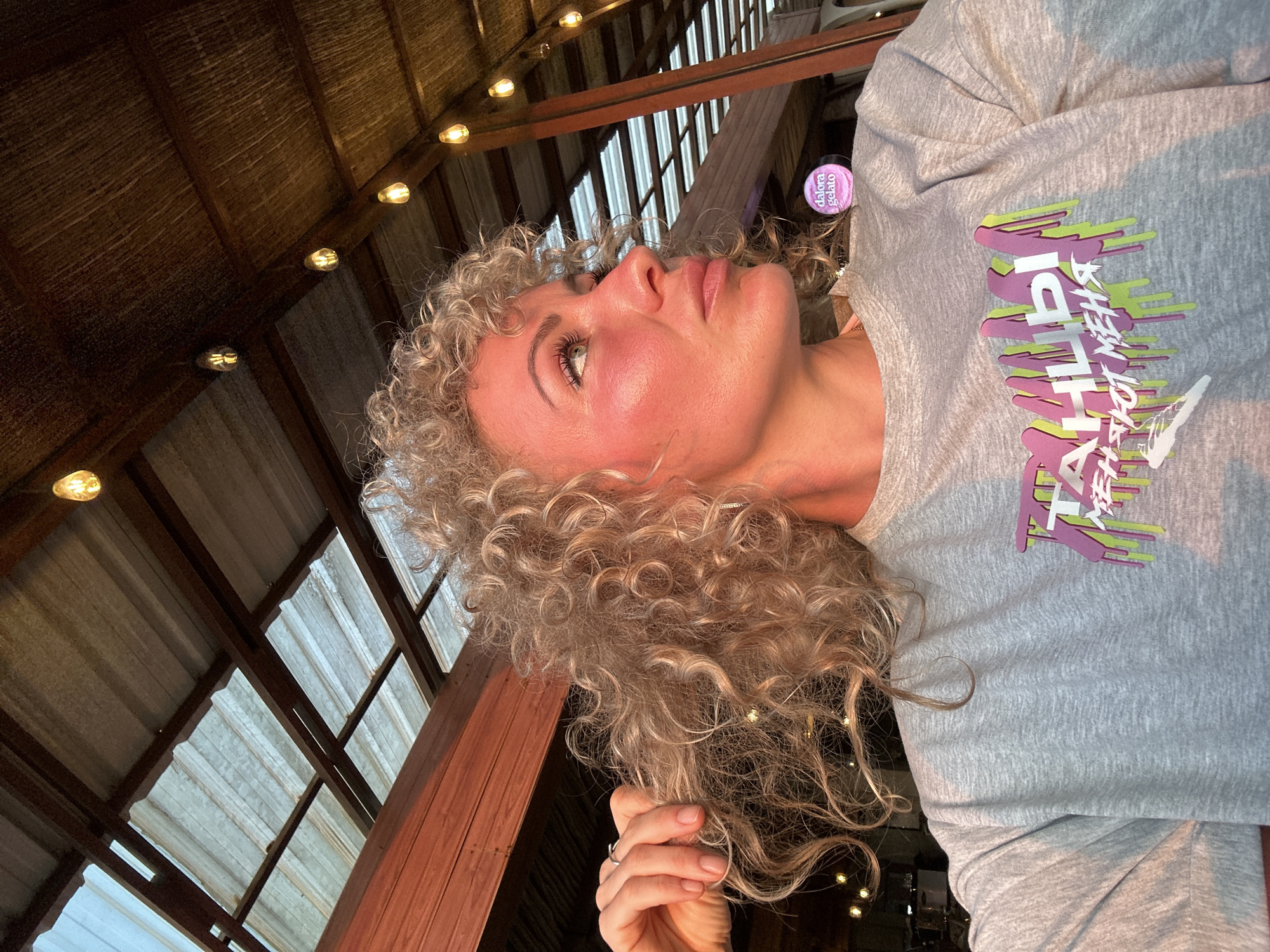 The image captures a woman with voluminous, light blonde curly hair, shown in a close-up profile view, looking upwards and slightly to the left. Her face is illuminated by a warm, golden light, highlighting her cheekbones and the bridge of her nose, with a subtle rosy blush. She wears a casual light grey long-sleeved t-shirt. The shirt features a prominent graphic design with a stylized, dripping effect in pink, lime green, and white. The text on the shirt includes a Cyrillic word, "ТАЙЛЫ," followed by "A NEW LOYALTY." A silver ring is visible on her right hand, which is gently holding a section of her curly hair.
The setting is an indoor space with a rustic architectural style. Above her, a ceiling structure is visible, composed of dark wooden beams and rafters, interspersed with what appears to be corrugated metal panels or skylights that allow some ambient light. Several warm-toned string lights are strung along the wooden beams, casting a soft, inviting glow across the scene. In the background, near the woman's right shoulder, a small, round, purple container with the text "dalora gelato" is partially visible. The overall atmosphere suggests a relaxed evening or a dimly lit indoor environment, possibly a restaurant, cafe, or a converted barn-like space.